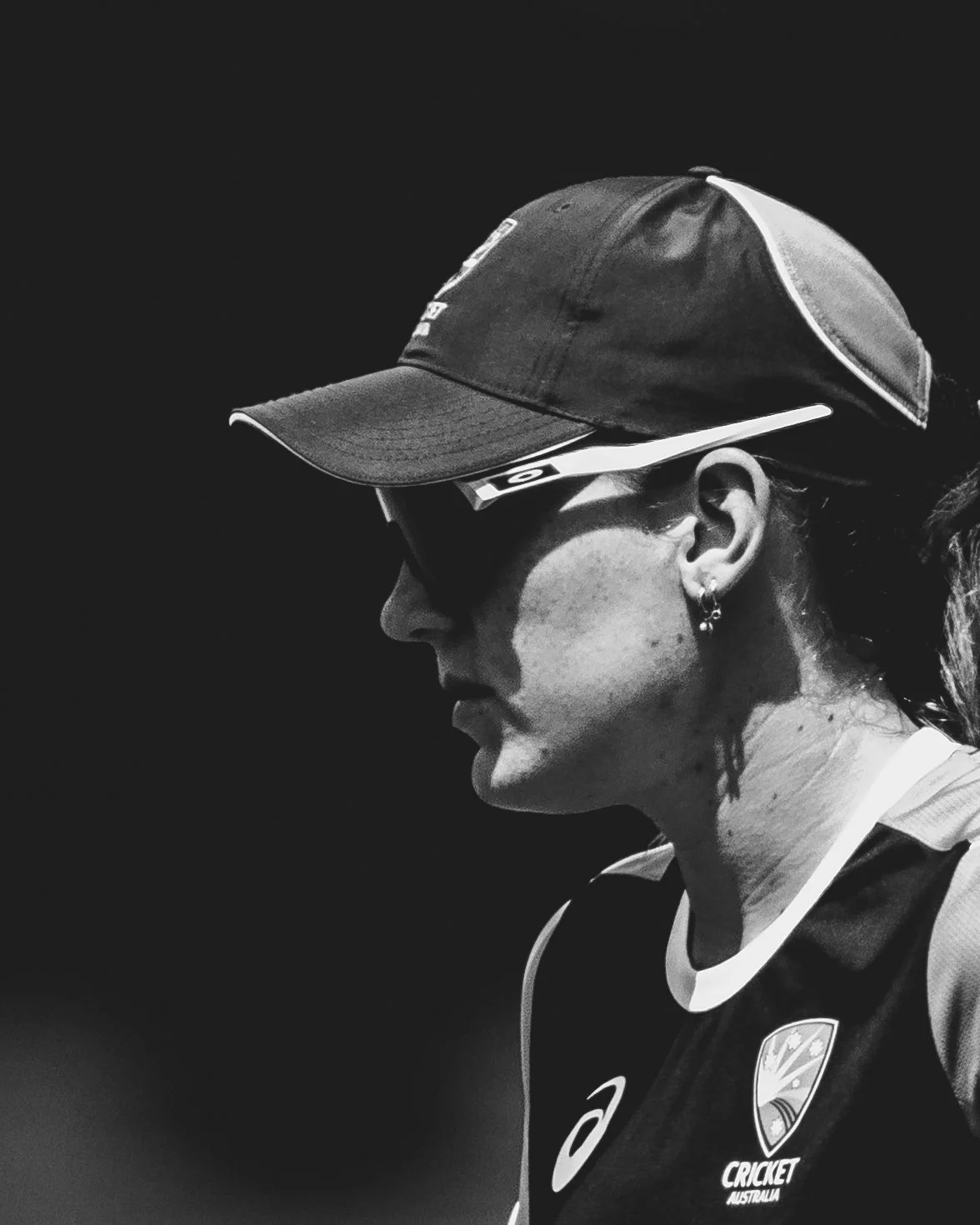 A female athlete in profile, wearing a cap, sunglasses, and a sports jersey with the Cricket Australia logo.