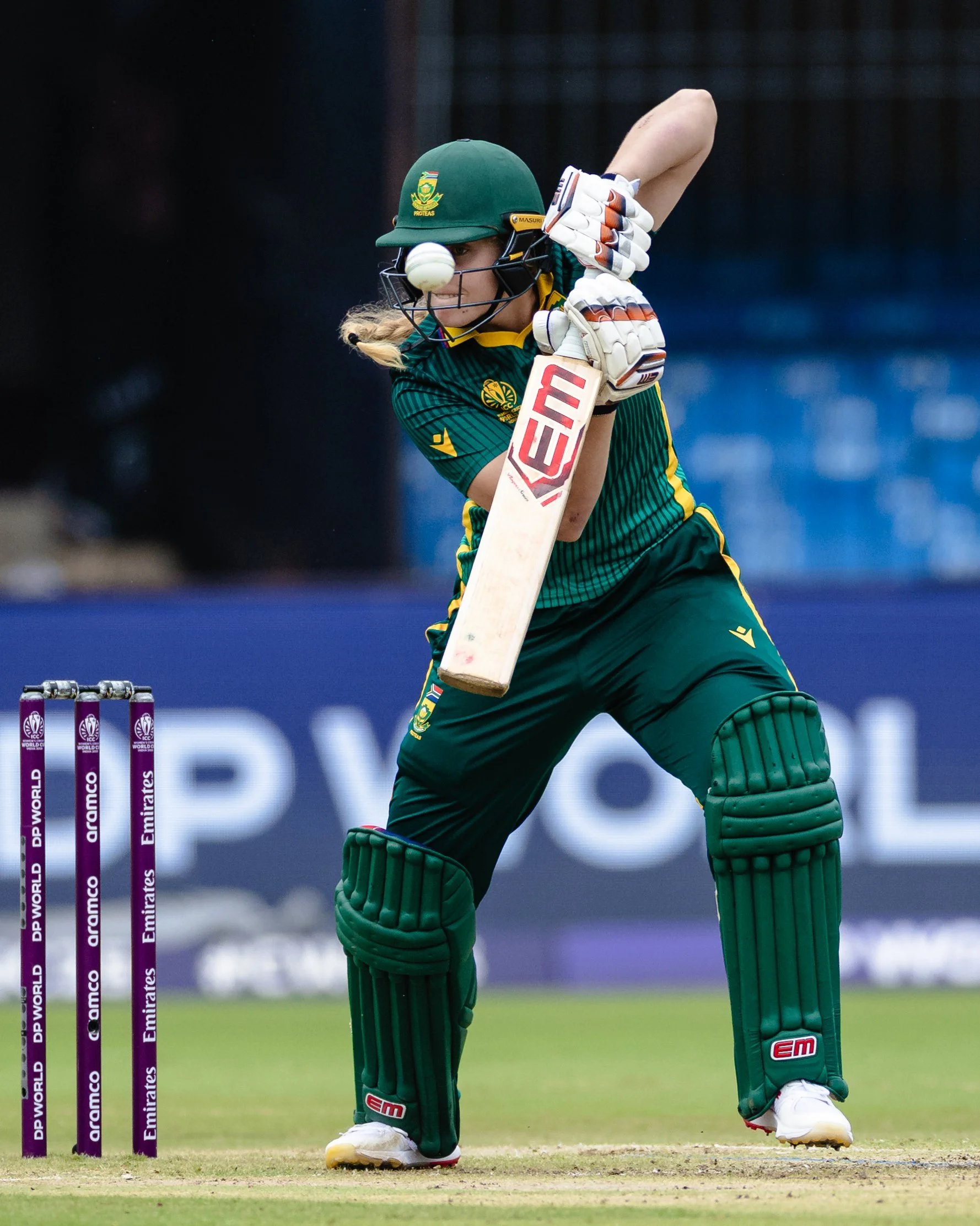 Cricketer in green uniform and batting helmet, trying to hit a cricket ball during a match, with purple advertising poles in the background.