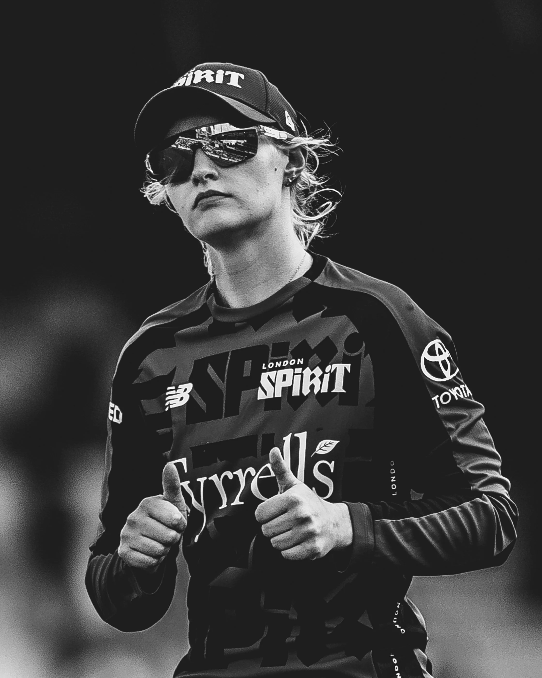 Black and white photo of a woman wearing a cap, sunglasses, and a sports jersey, giving a thumbs-up.