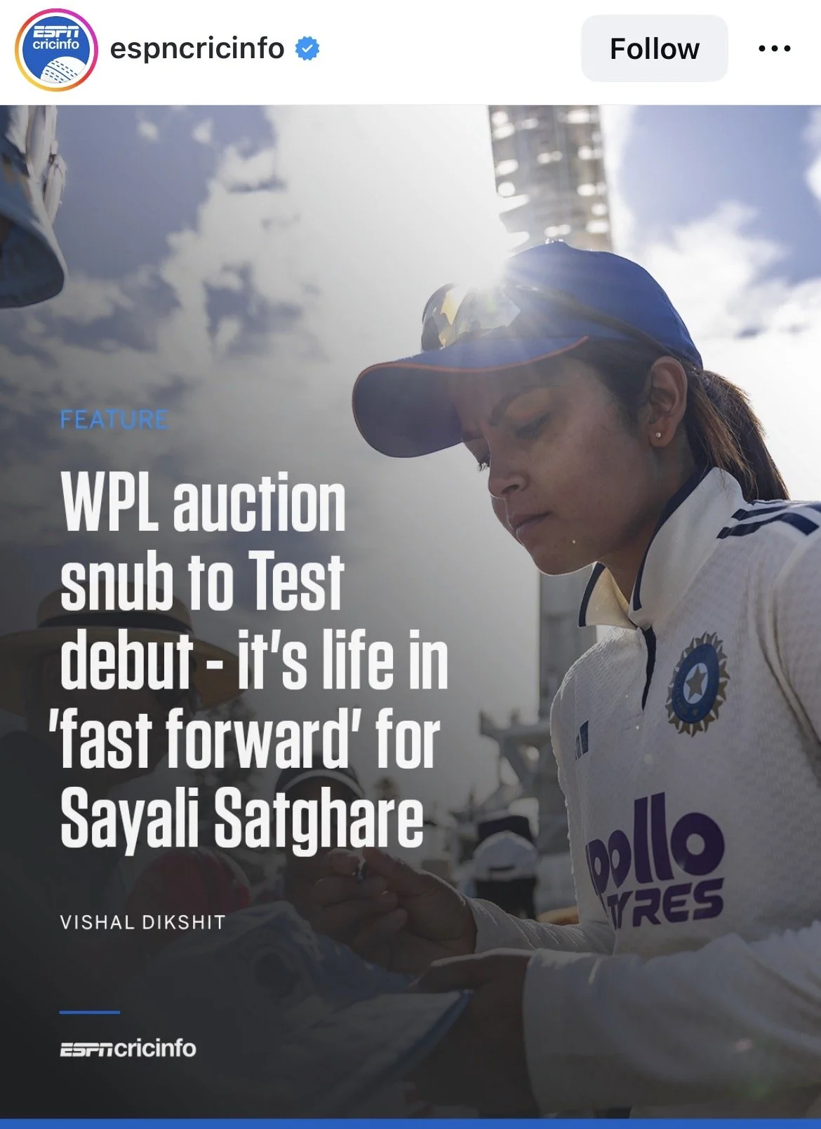 A female cricketer wearing a blue cap and white jersey with the Indian cricket team logo, looking downwards with sunlight and clouds in the background, during a cricket event.