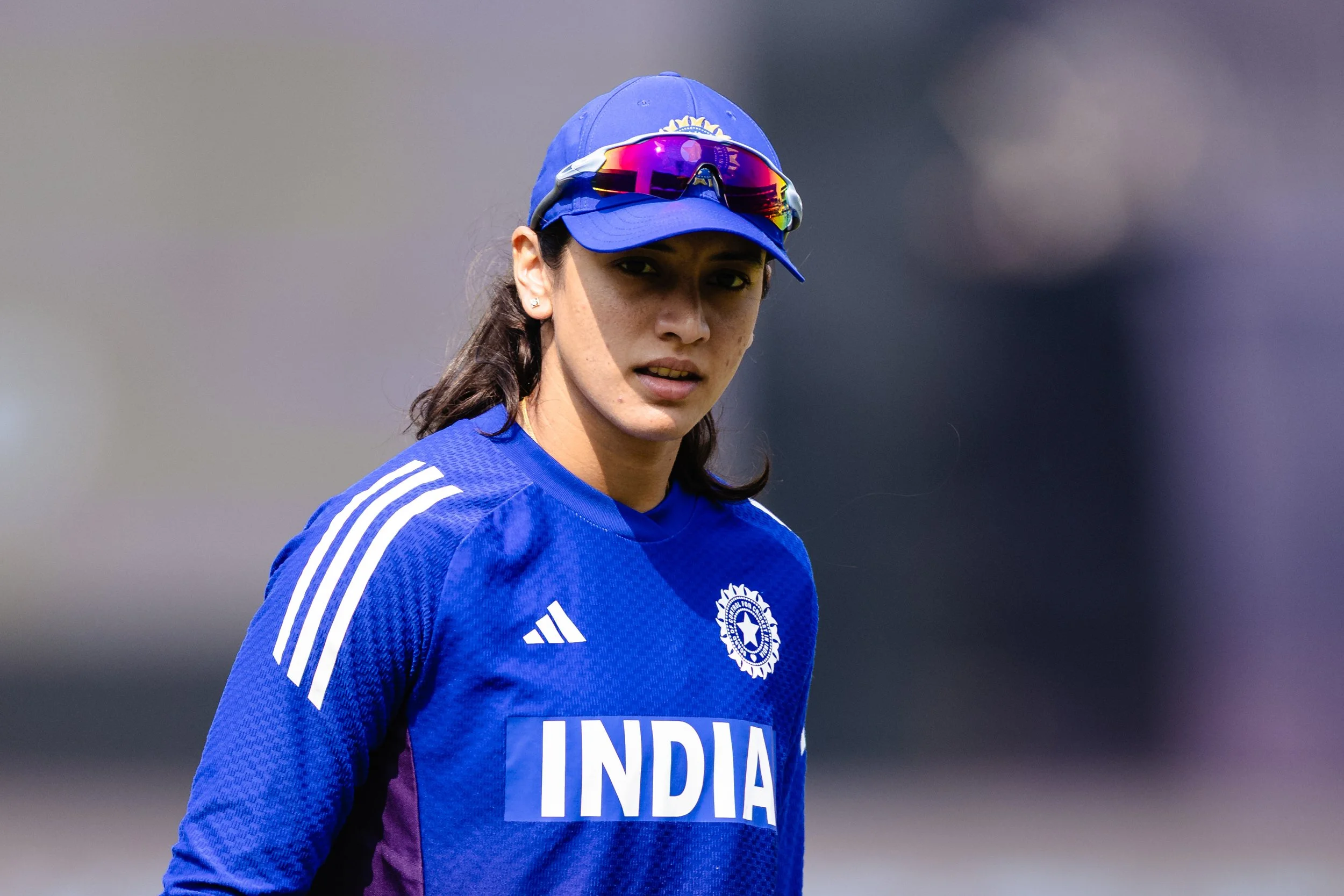 A woman wearing a blue sports jersey with 'INDIA' written on it, a blue cap, and sports sunglasses, during a cricket match.