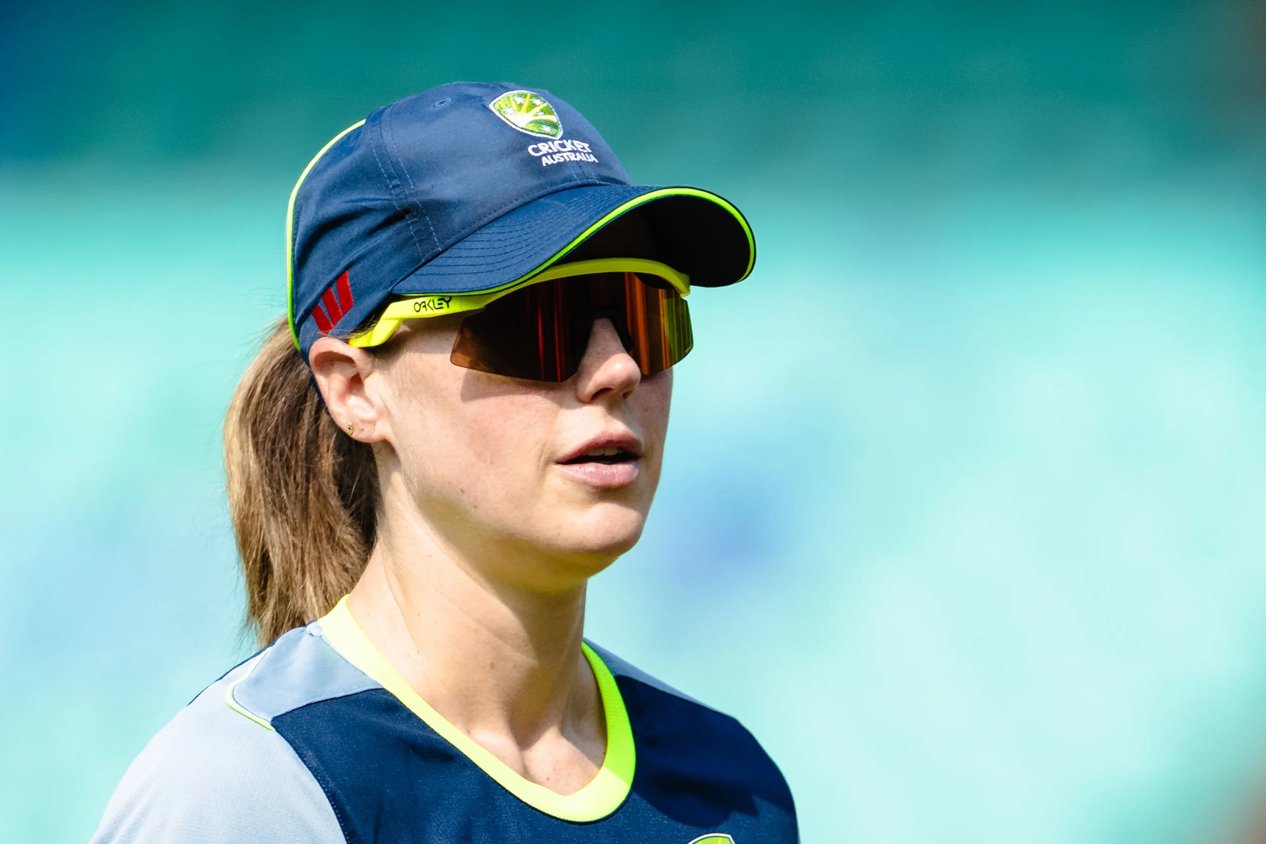 A woman wearing a navy blue cricket hat with an Australia cricket logo, sports sunglasses, and a navy blue sporting jersey against a blurry blue-green background.