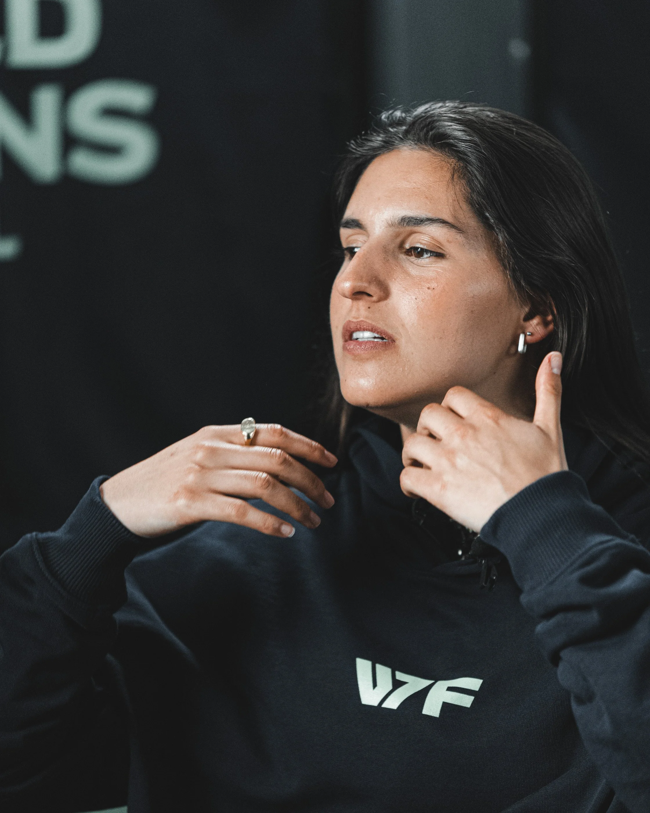 Close-up of a woman with dark hair, wearing earrings and a black hoodie, touching her neck and chin, with a dark background and partially visible text on her hoodie.