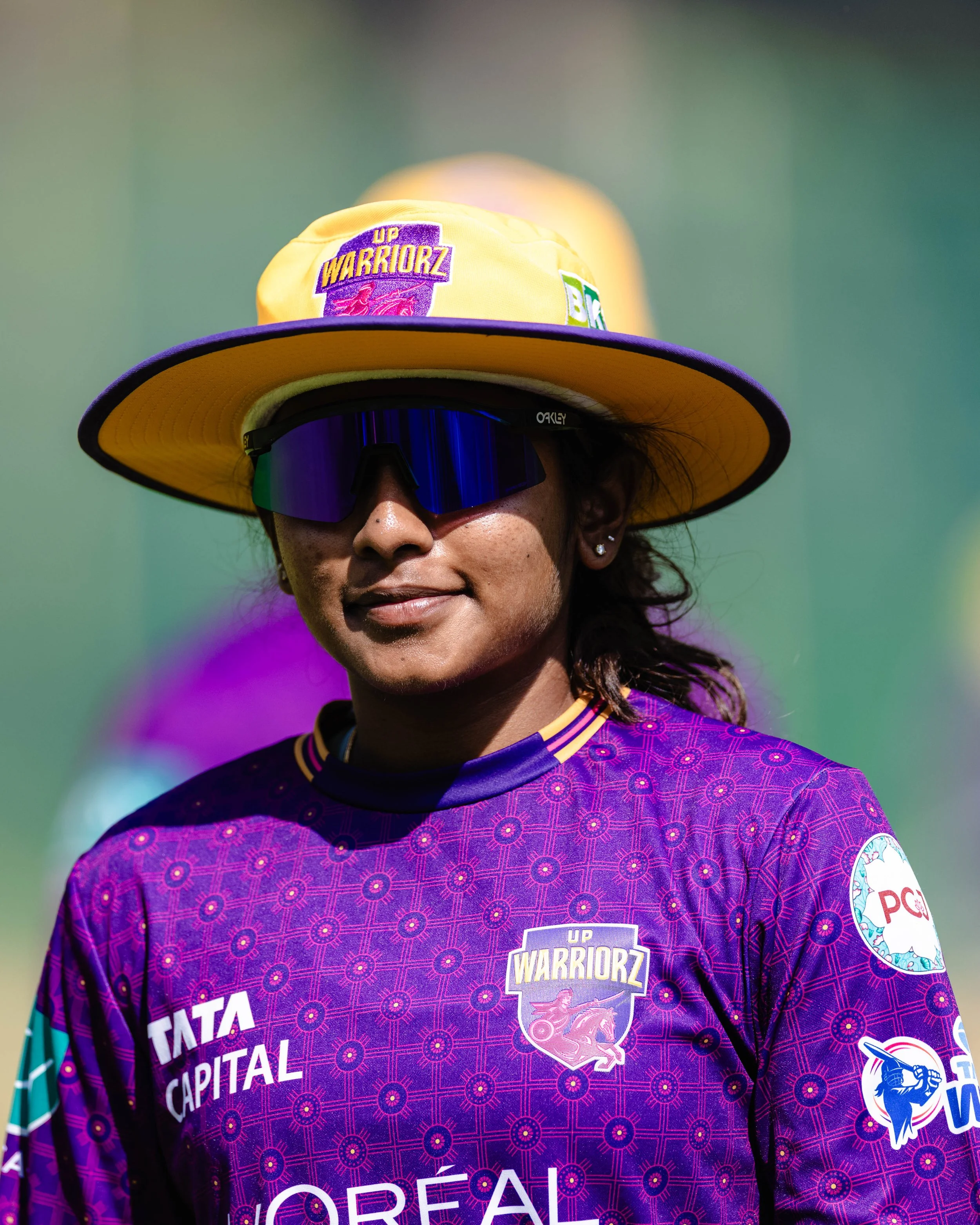 A woman wearing a wide yellow hat with purple trim, sunglasses, and a purple sports jersey with various logos and patches, participating in a sports event.