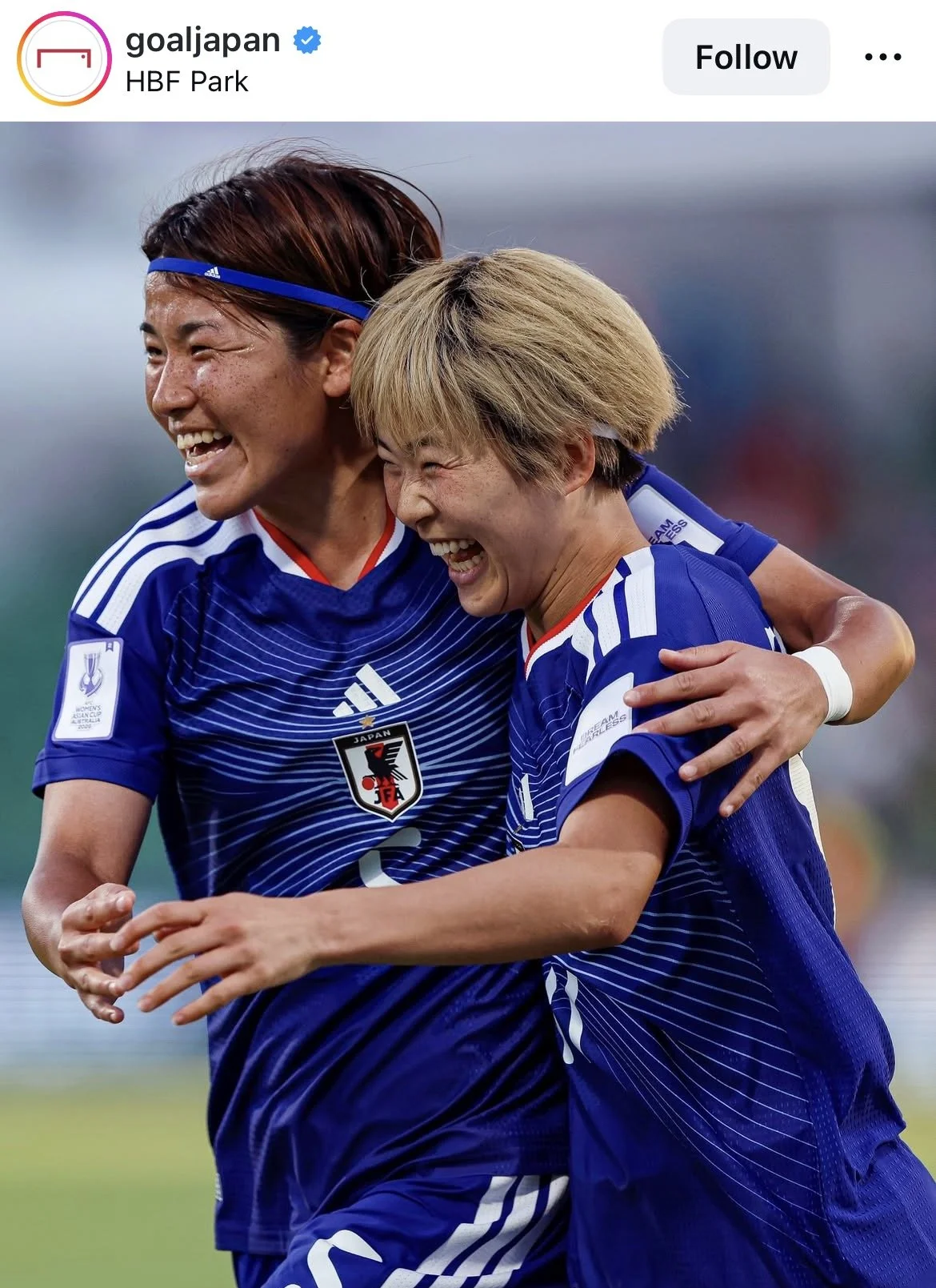 Two female soccer players in blue jerseys celebrating and hugging each other on the field, smiling joyfully.