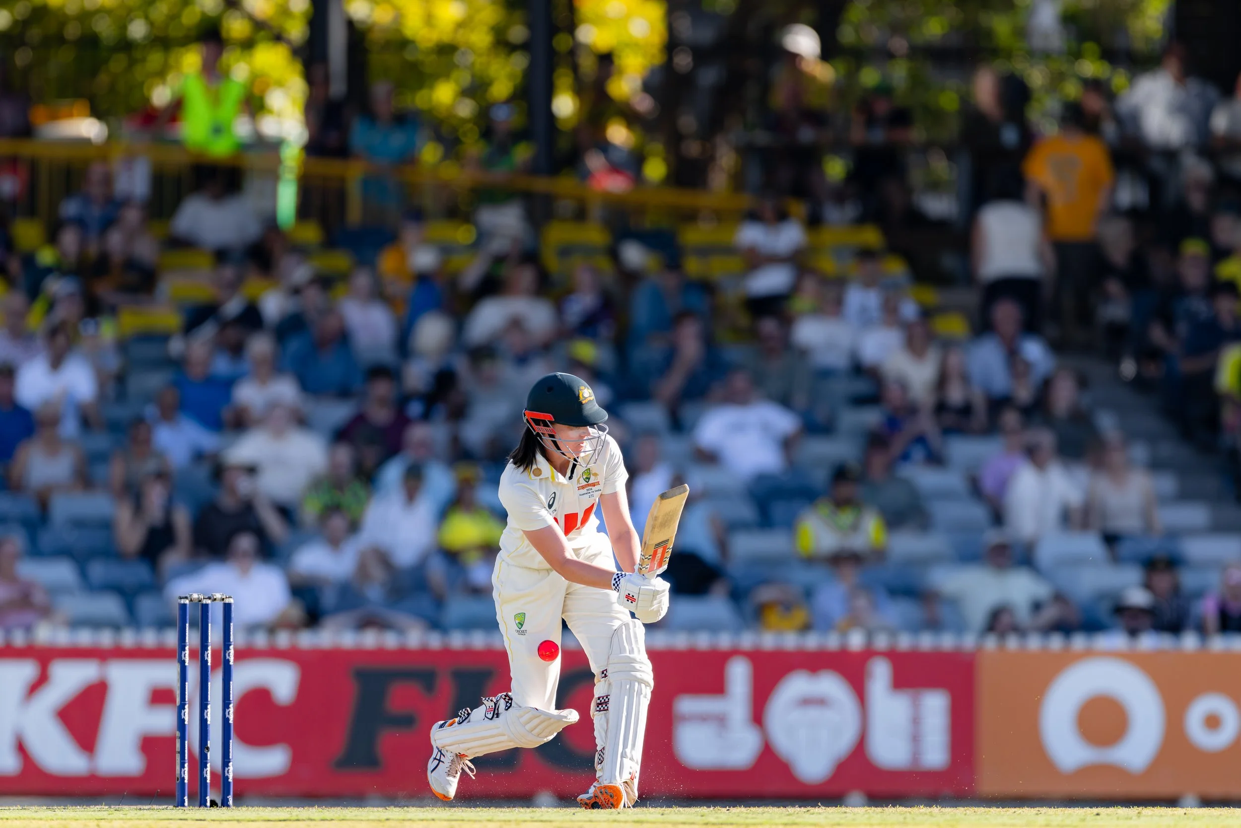 Cricket player wearing white uniform and black helmet batting on the field during a game, with a blurred audience in the background.