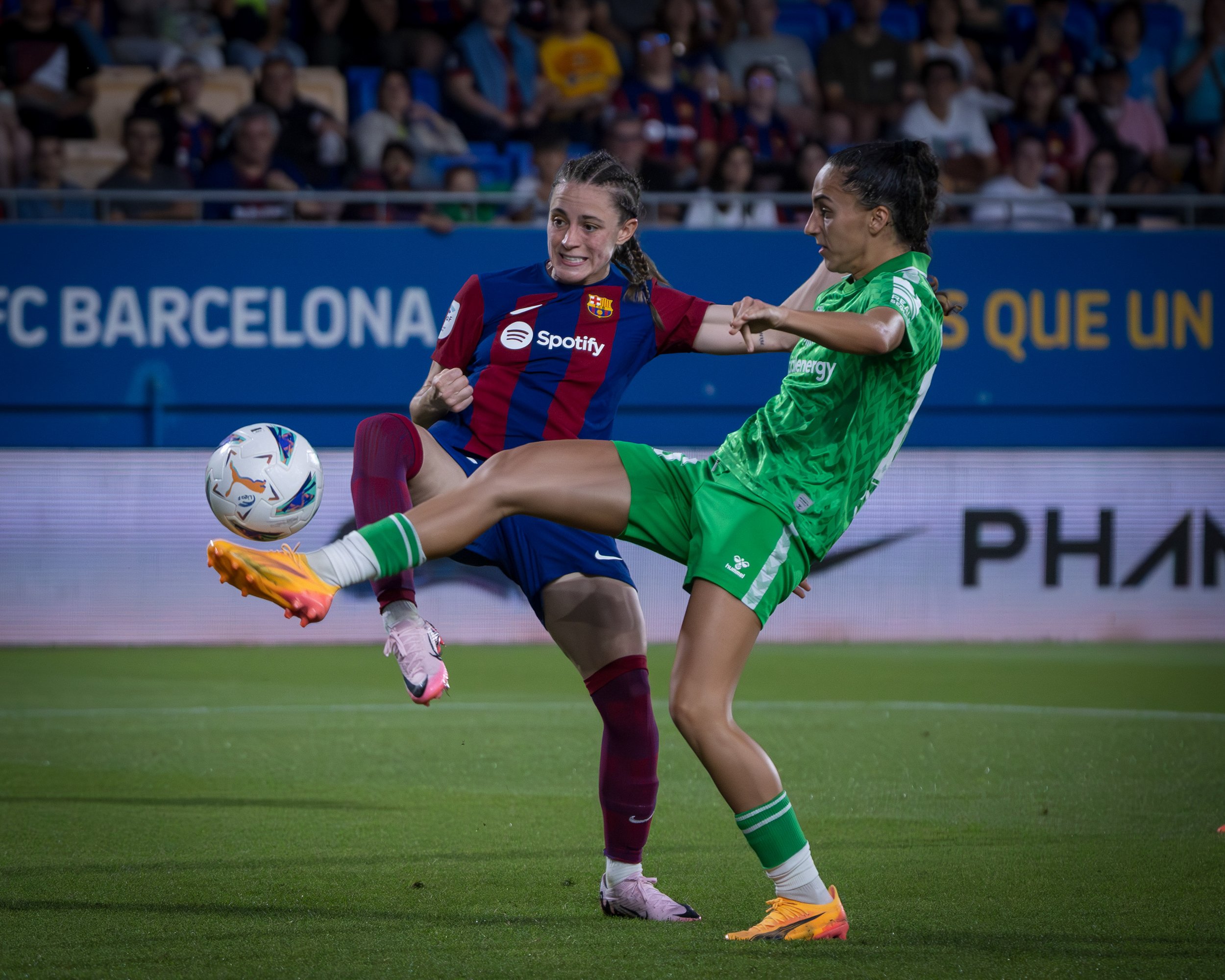 Two female soccer players competing for the ball during a match, with a crowd in the background.