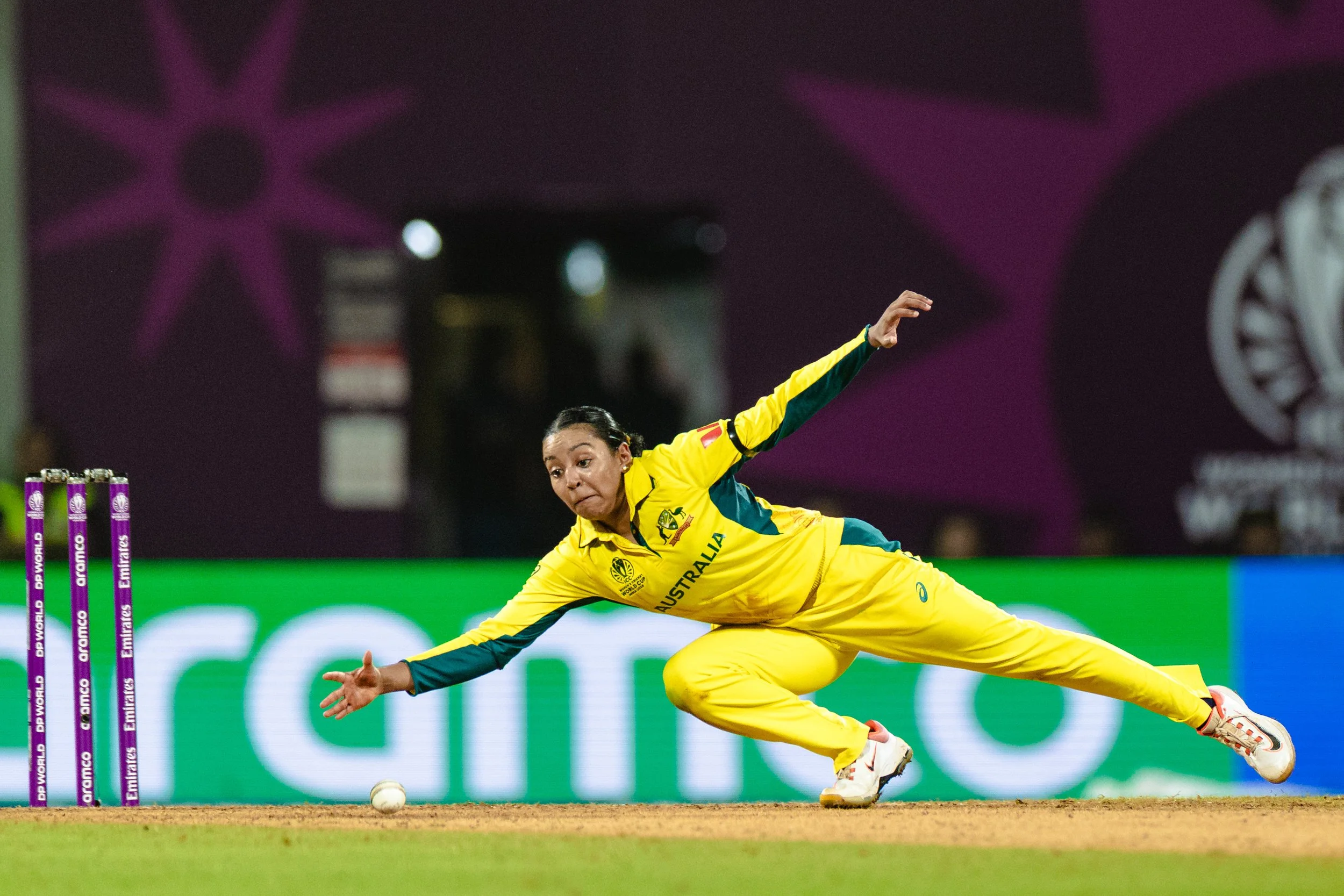 A female cricket player from Australia in a yellow uniform diving to catch the ball near the stumps during a match.