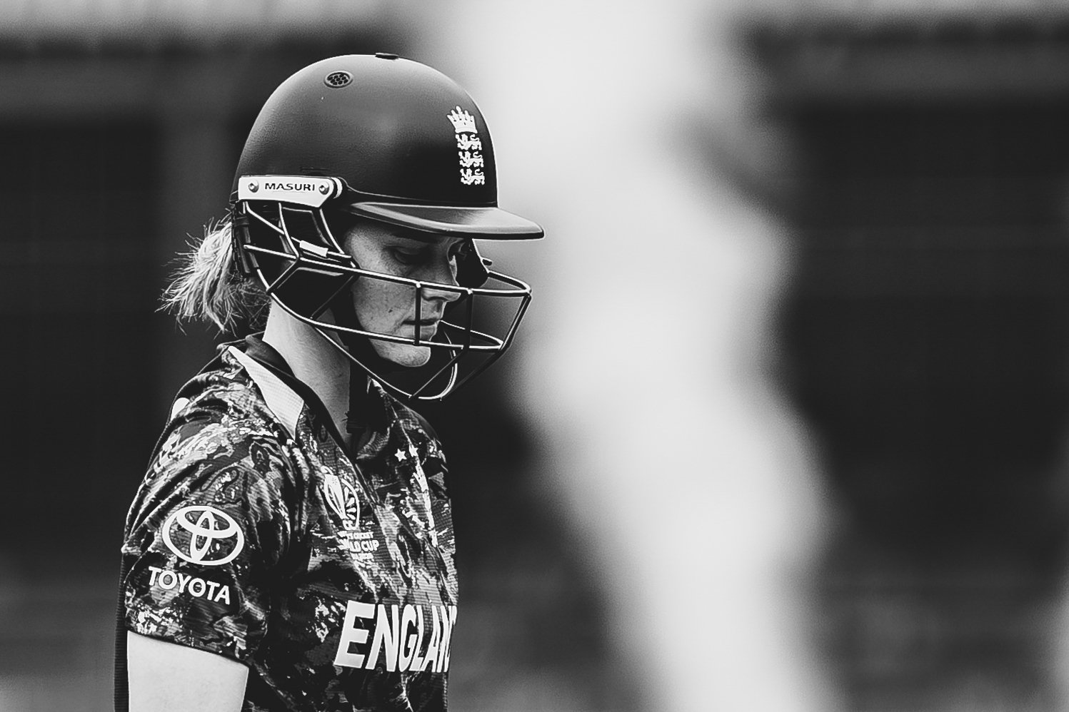 A female cricket player wearing a helmet and sports jersey with sponsor logos, looking down with a focused expression.