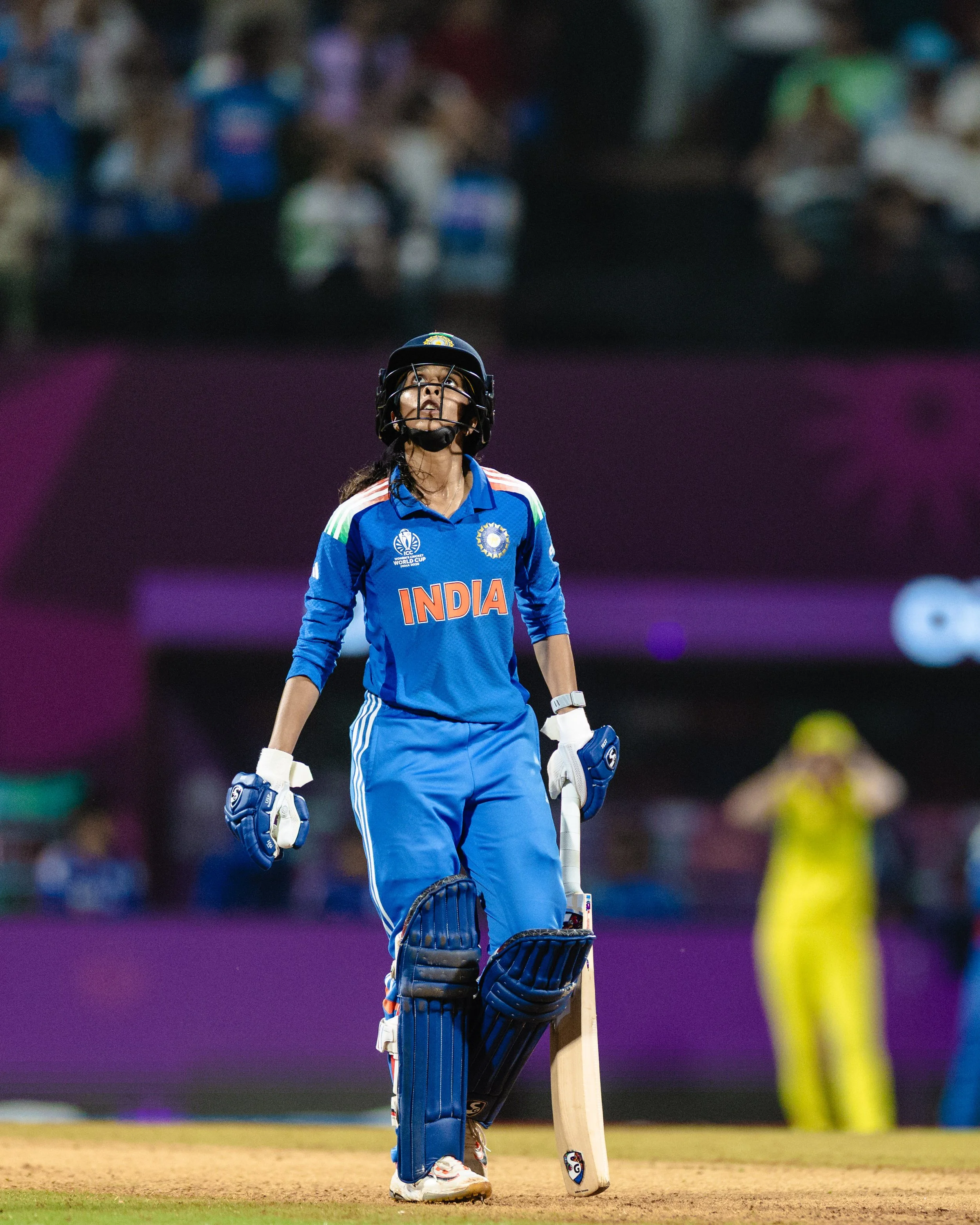 A female Indian cricket player on the field in blue uniform, wearing a helmet and gloves, holding a cricket bat, looking upward during a match.