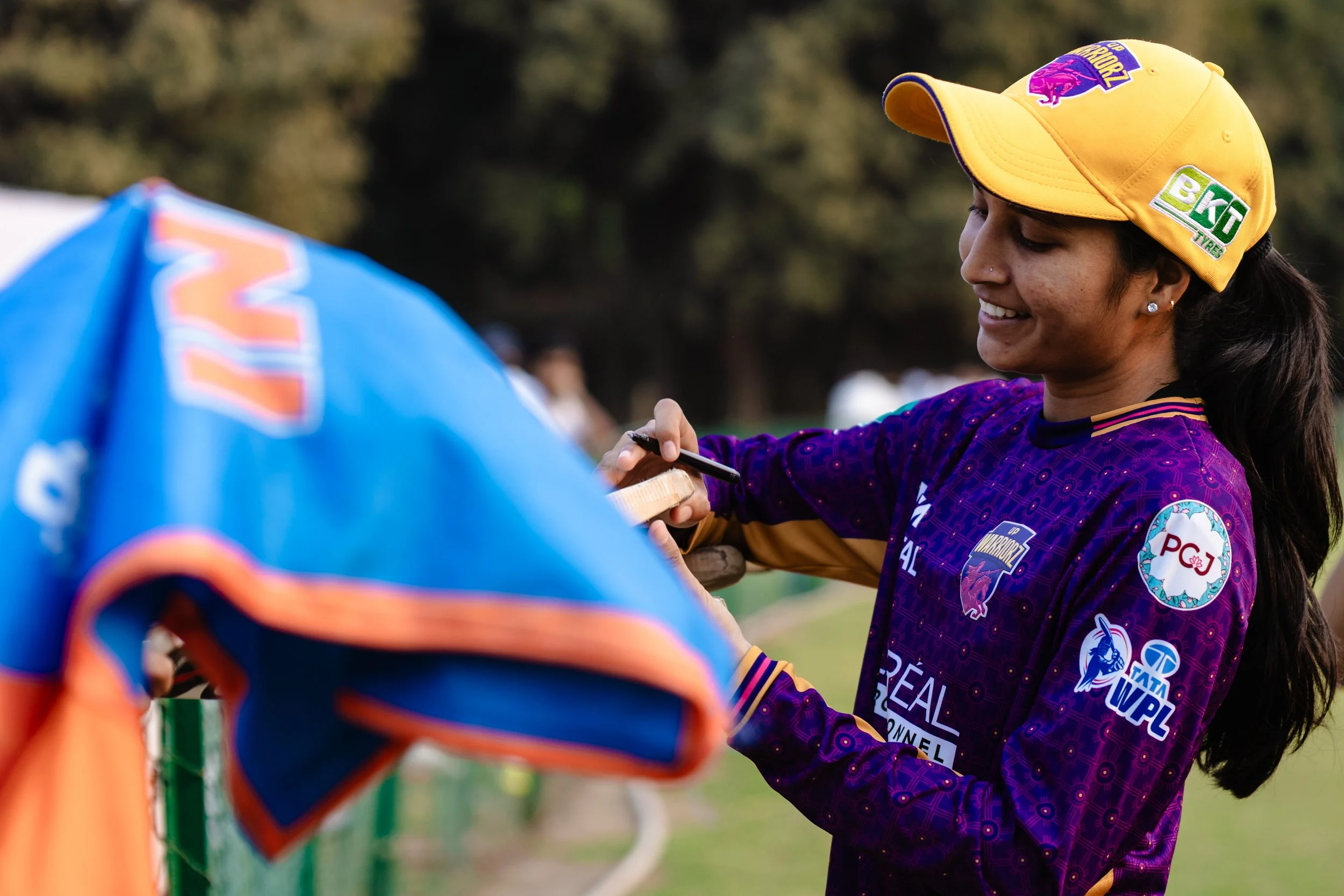 A woman wearing a yellow cap and a purple sports jersey is smiling while signing an object, possibly a helmet, during an outdoor sports event with a green natural background.