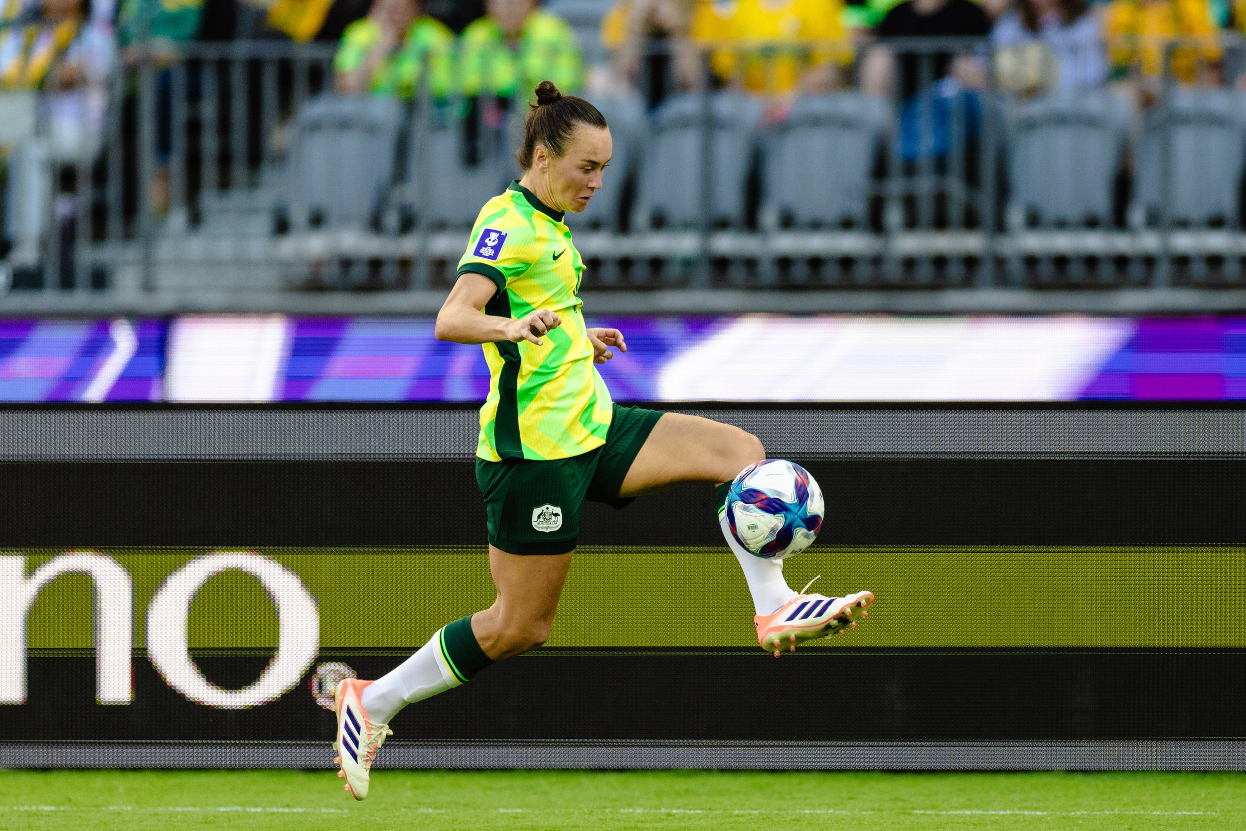 A female soccer player in a bright yellow and green jersey, black shorts, and white and pink cleats, is captured mid-step on the soccer field during a game, with an audience and stadium seating in the background.