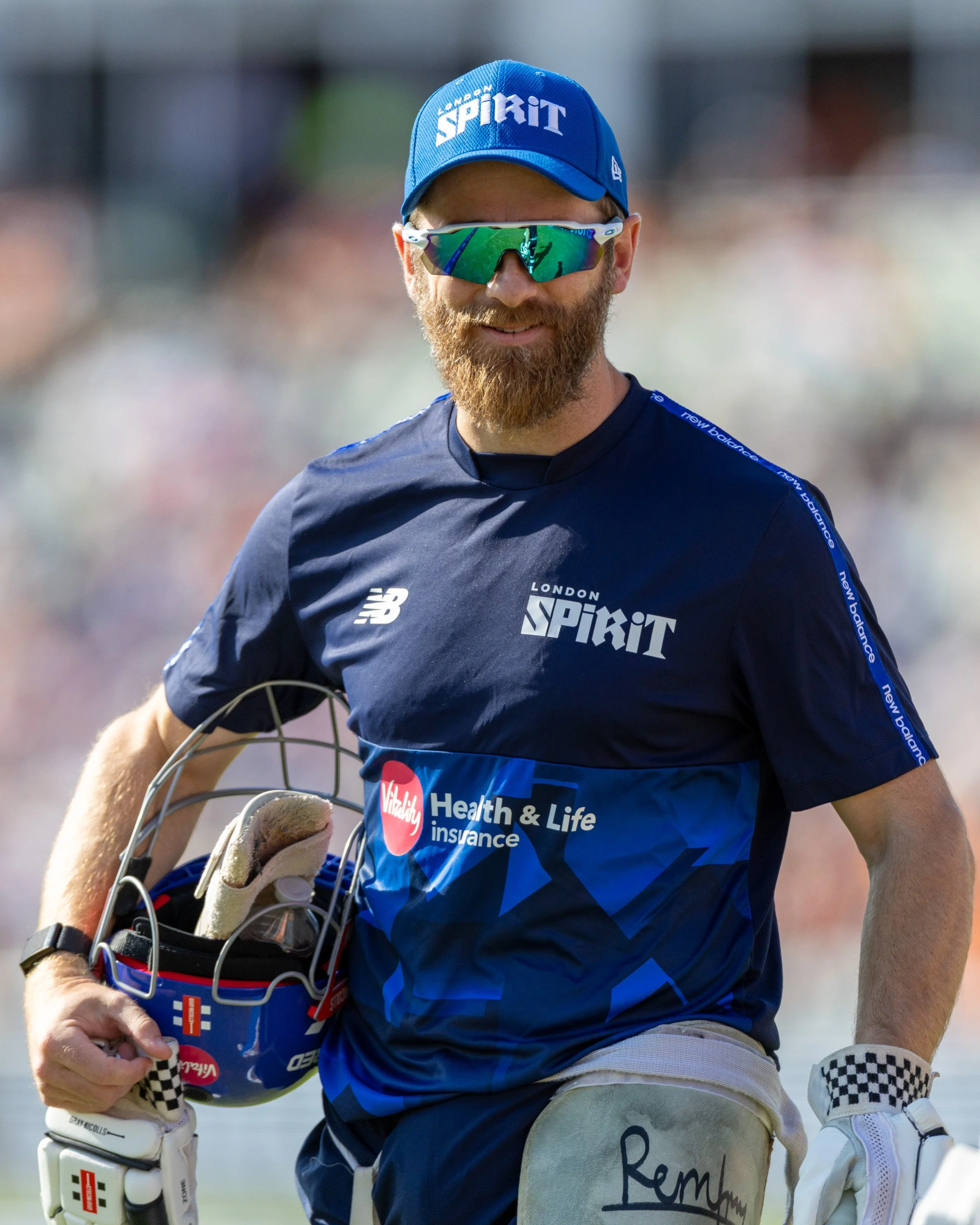 A man with a beard running on a sports field. He is wearing sunglasses, a blue cap with the words 'London Spirit,' a dark blue sports shirt with 'London Spirit' logo, and rugby gloves. He is carrying a rugby ball.