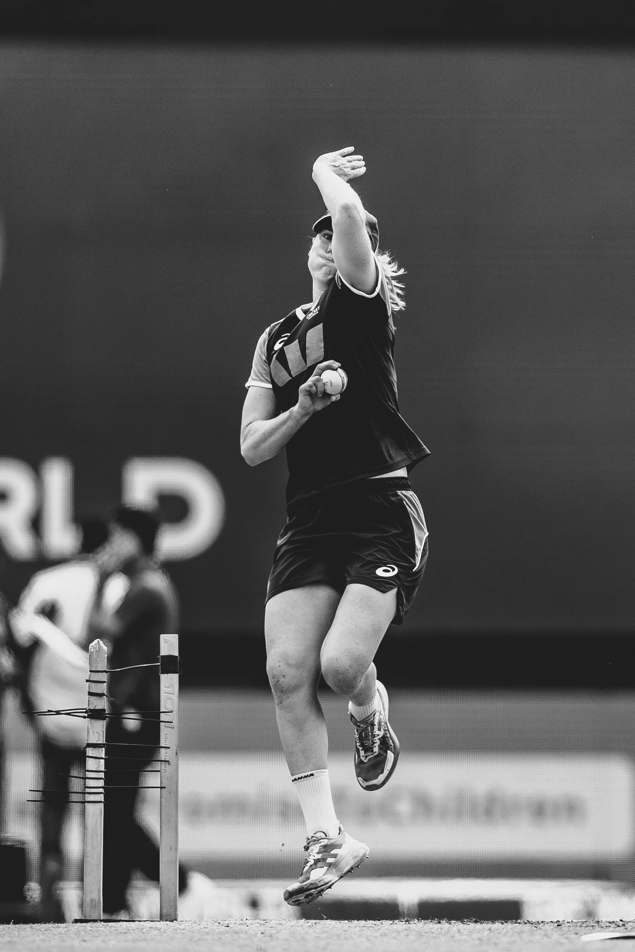 Black and white photo of a female athlete jumping over hurdles during a track and field event. She is wearing a sports uniform, a cap, and running shoes, with a focused expression.
