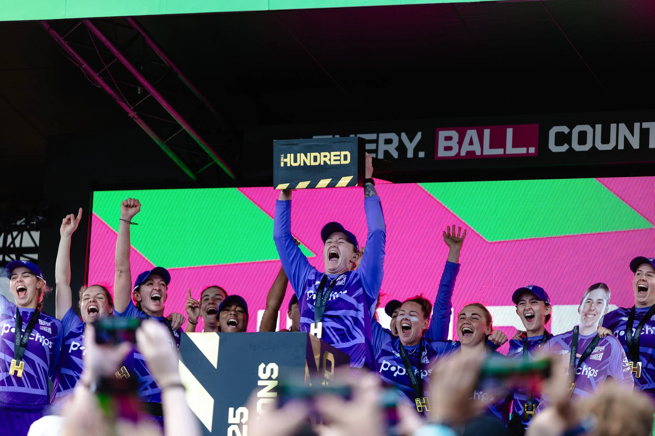 Group of women celebrating on a podium with medals, one woman in the center holds a sign that reads 'The Hundred,' in a sports event setting.