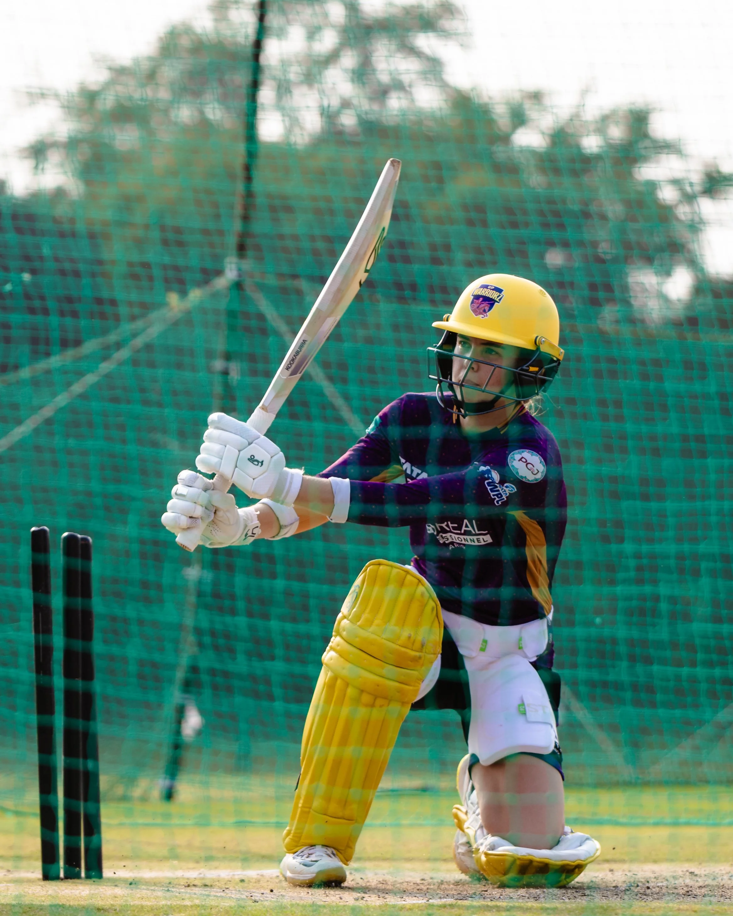 A cricket goalkeeper wearing a yellow helmet, purple jersey, and yellow leg pads, holding a cricket bat in front of a wicket with a green net in the background.