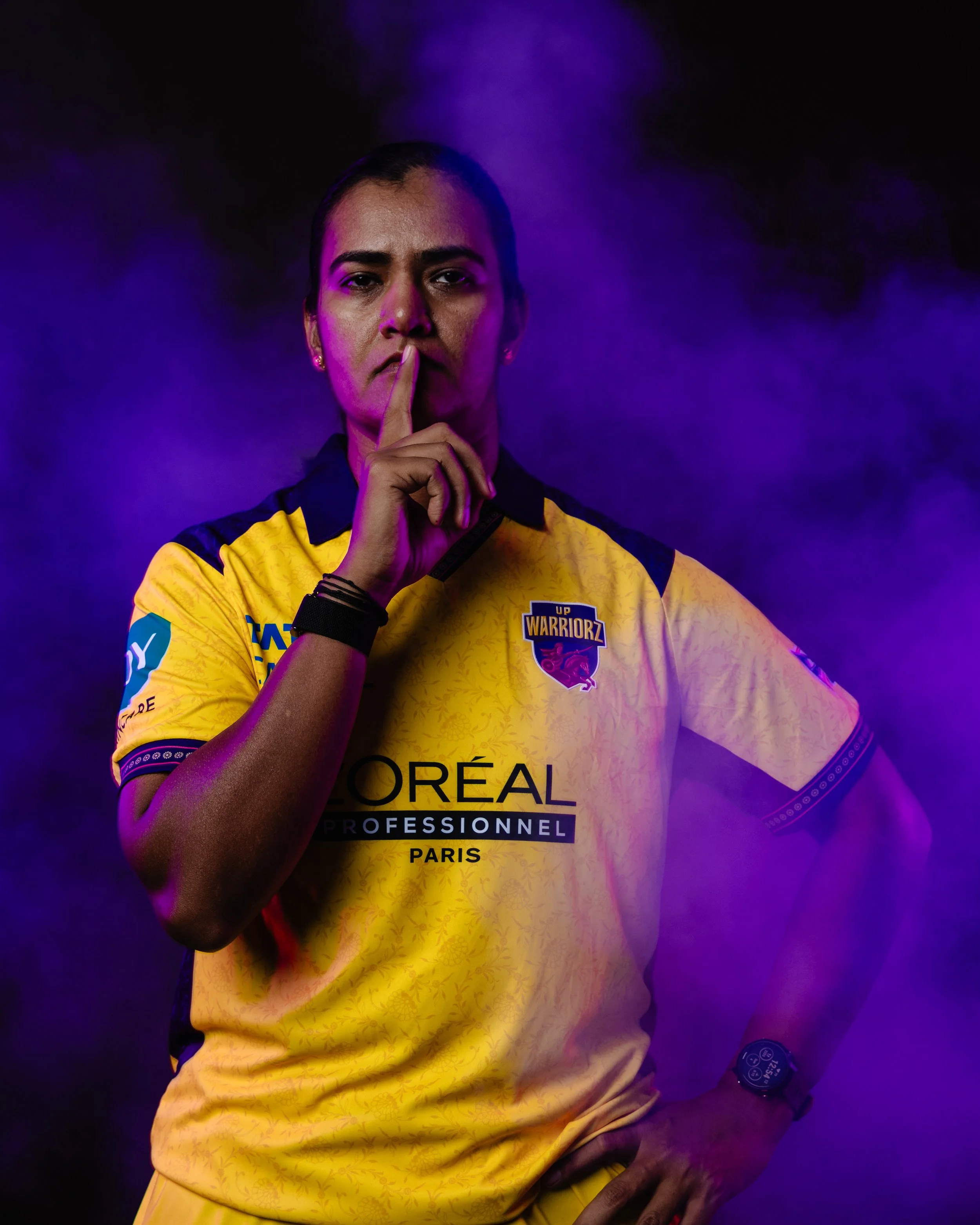 A woman in a yellow sports jersey with the logo of UP Warriorz, an Indian Premier League cricket team, raising her index finger to her lips in a gesture of silence against a dark purple background.