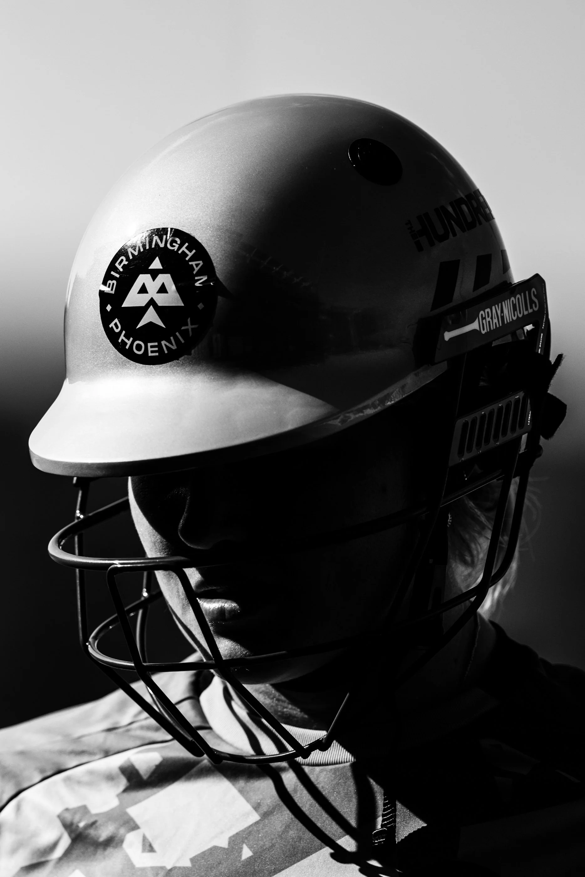 A black and white photograph of a football helmet worn by a person with a face mask. The helmet has a logo that reads 'Birmingham Phoenix' with a stylized phoenix symbol. There are some words on the side of the helmet, including 'HUNDER' and 'GRAY NI