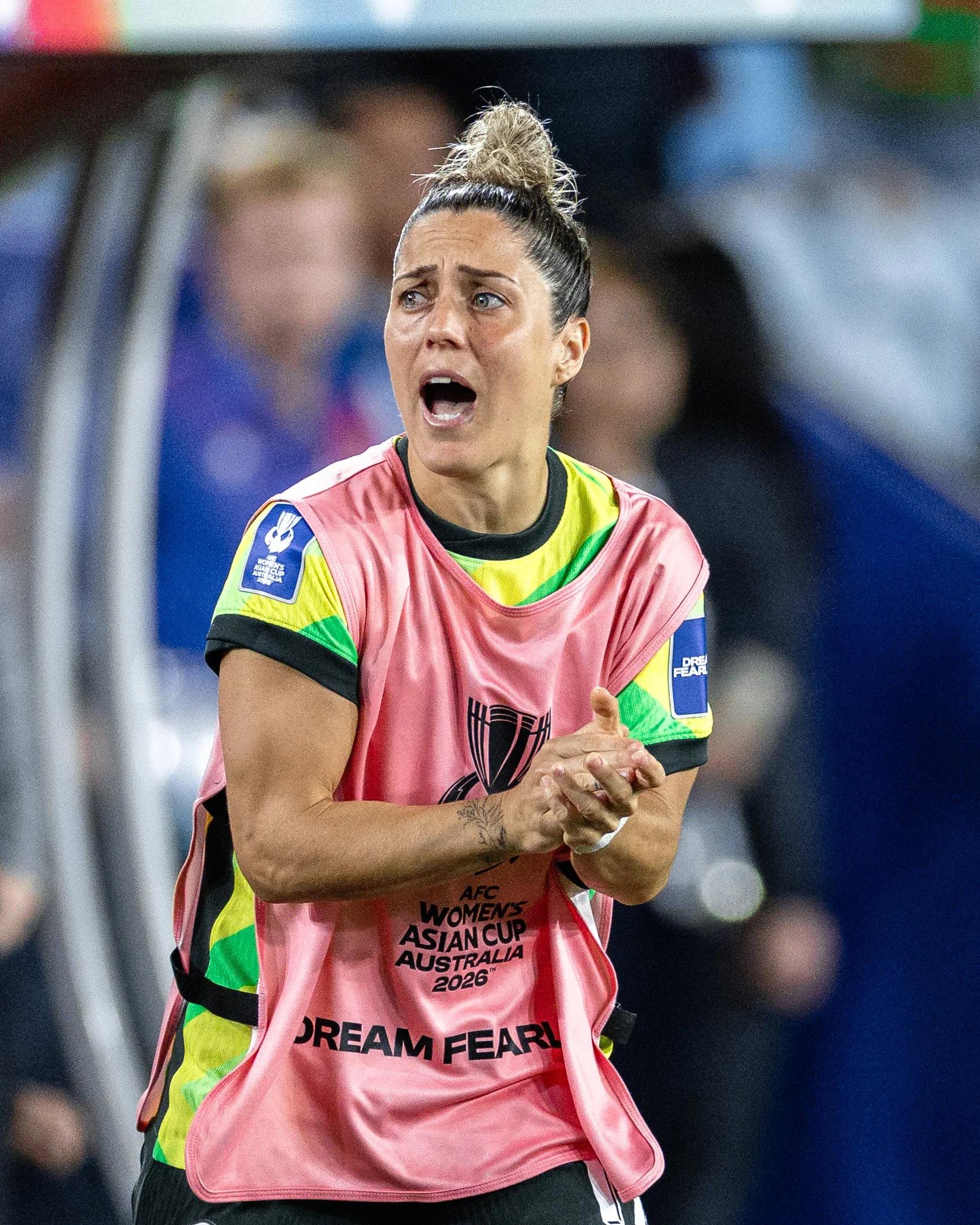 Female soccer player with dark hair in a bun, wearing a pink vest with text, and clapping her hands during a game.