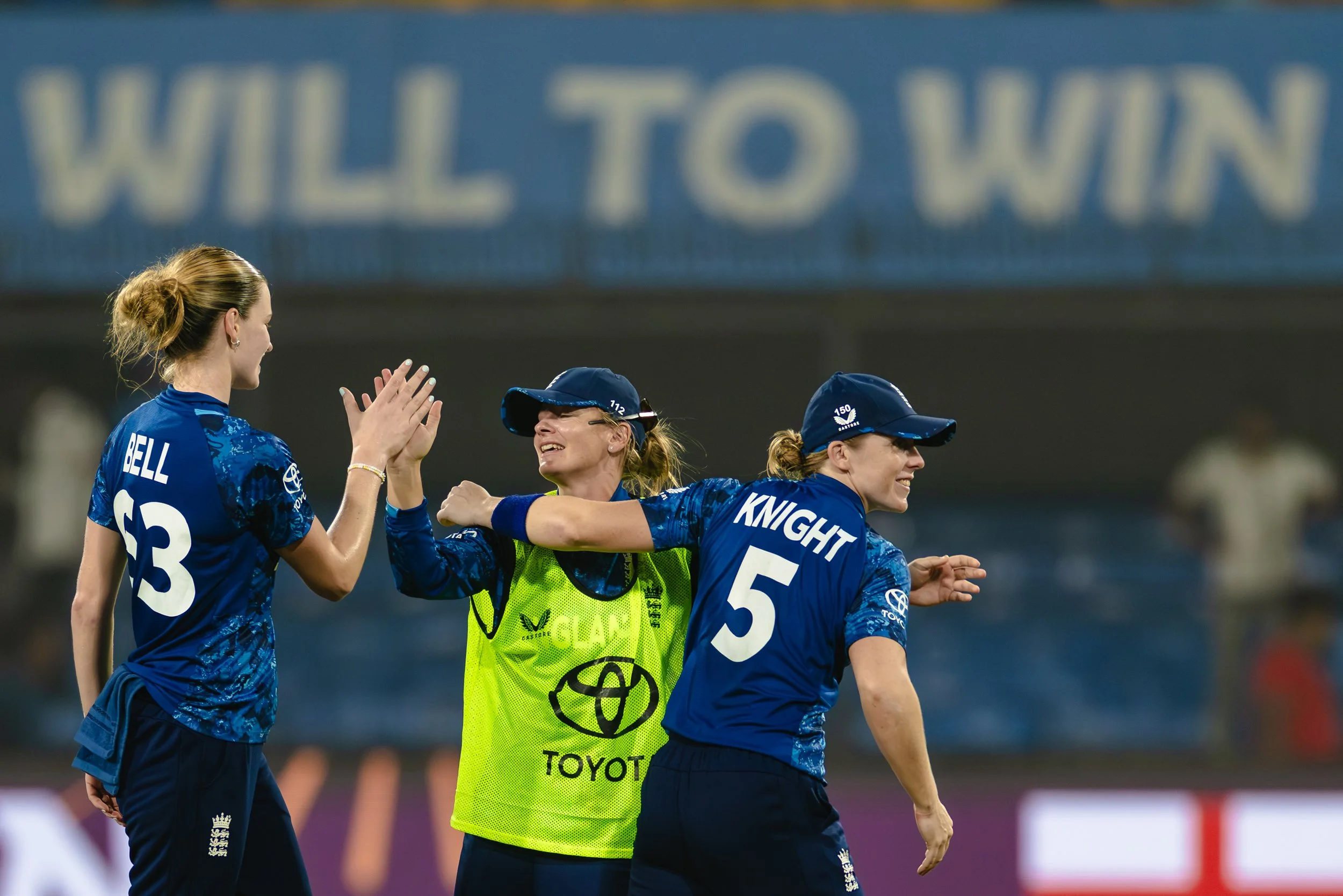 Three female soccer players celebrating on the field, two wearing blue jerseys with numbers 13 and 5, and one wearing a yellow vest with the word 'GLAM' and a Toyota logo, against a blurred stadium background.