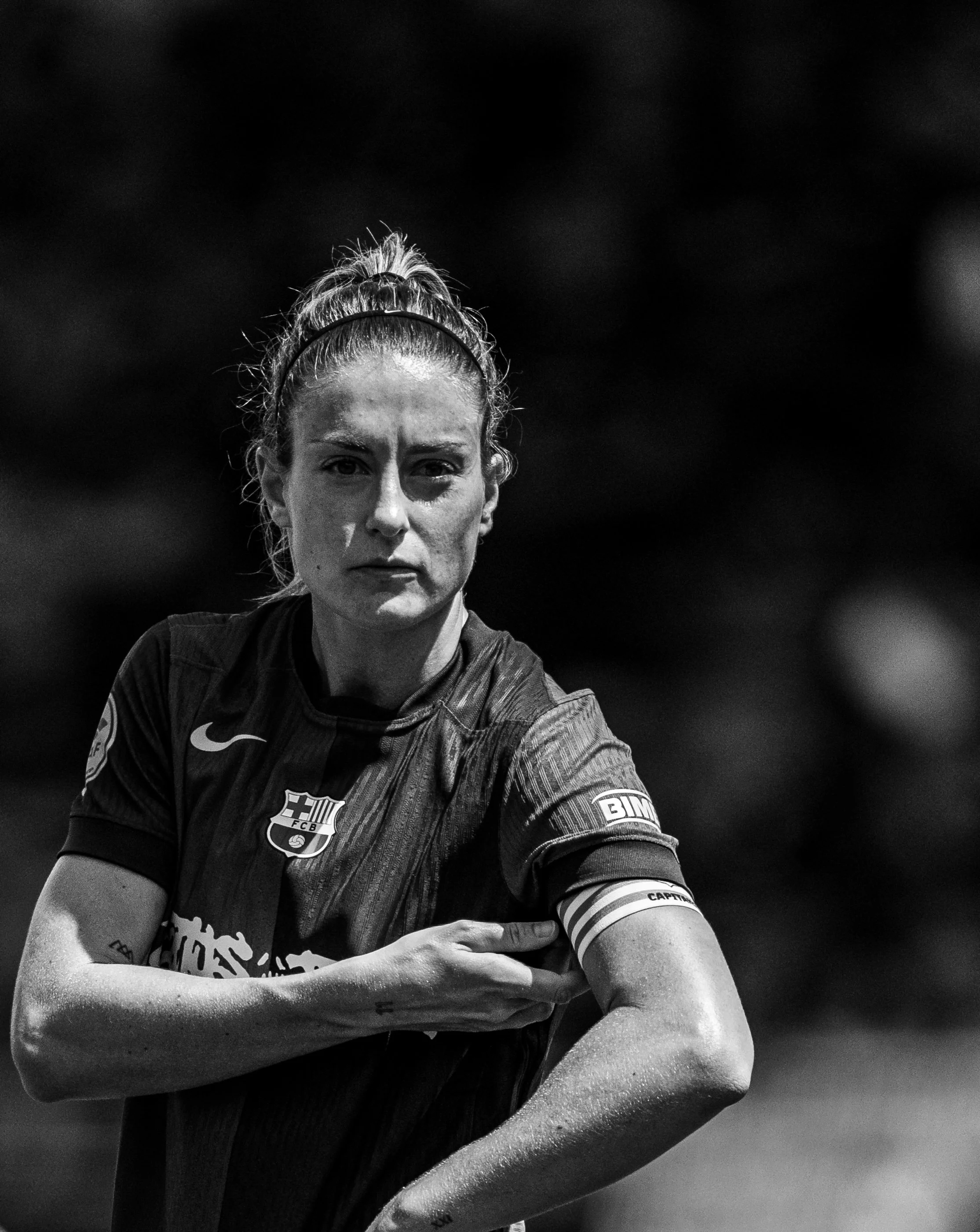 A female soccer player with a serious expression, wearing a jersey with FC Barcelona's logo and captain's armband, standing with crossed arms.