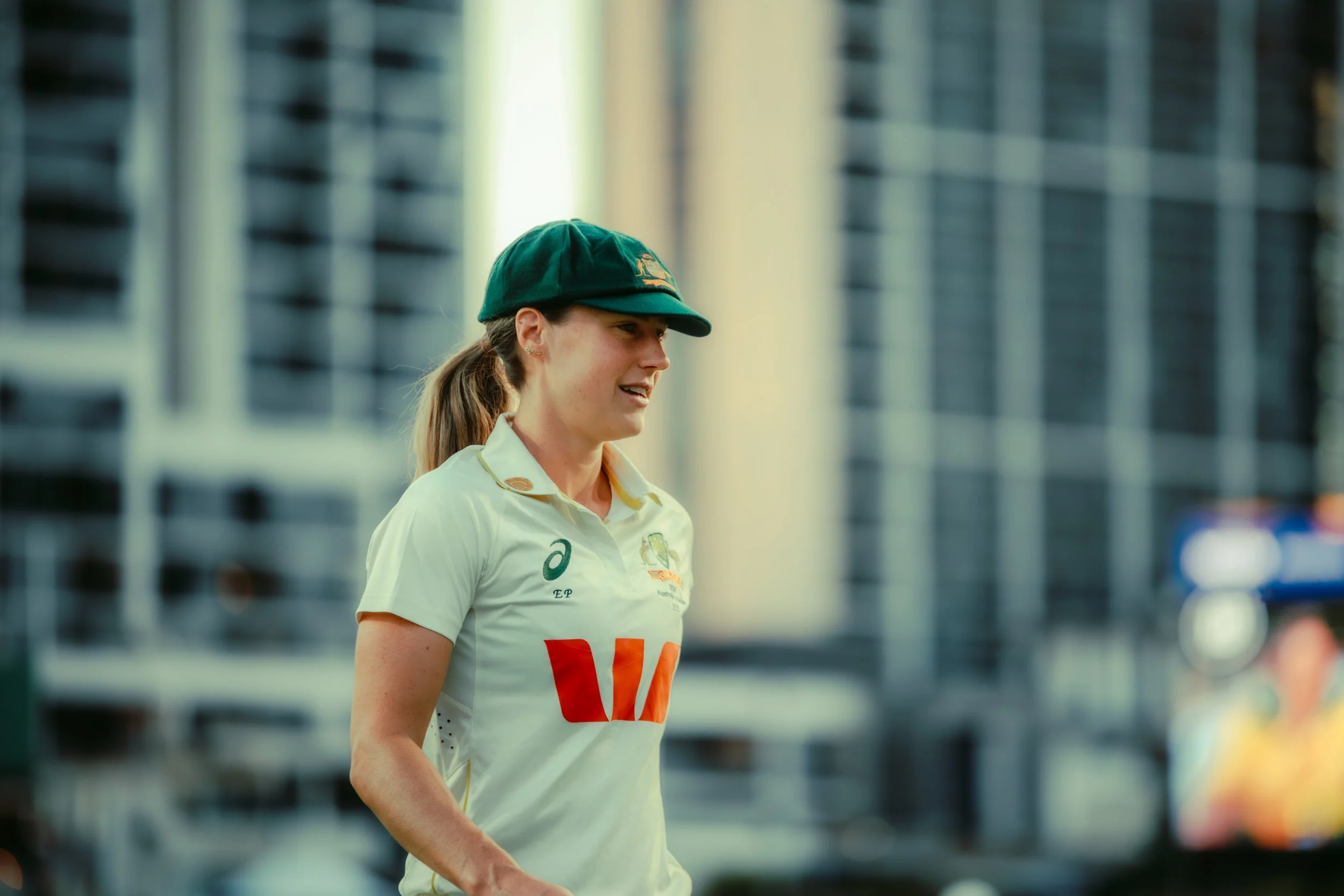 A female athlete in a white sports uniform and green cap, walking outdoors in front of blurred buildings.