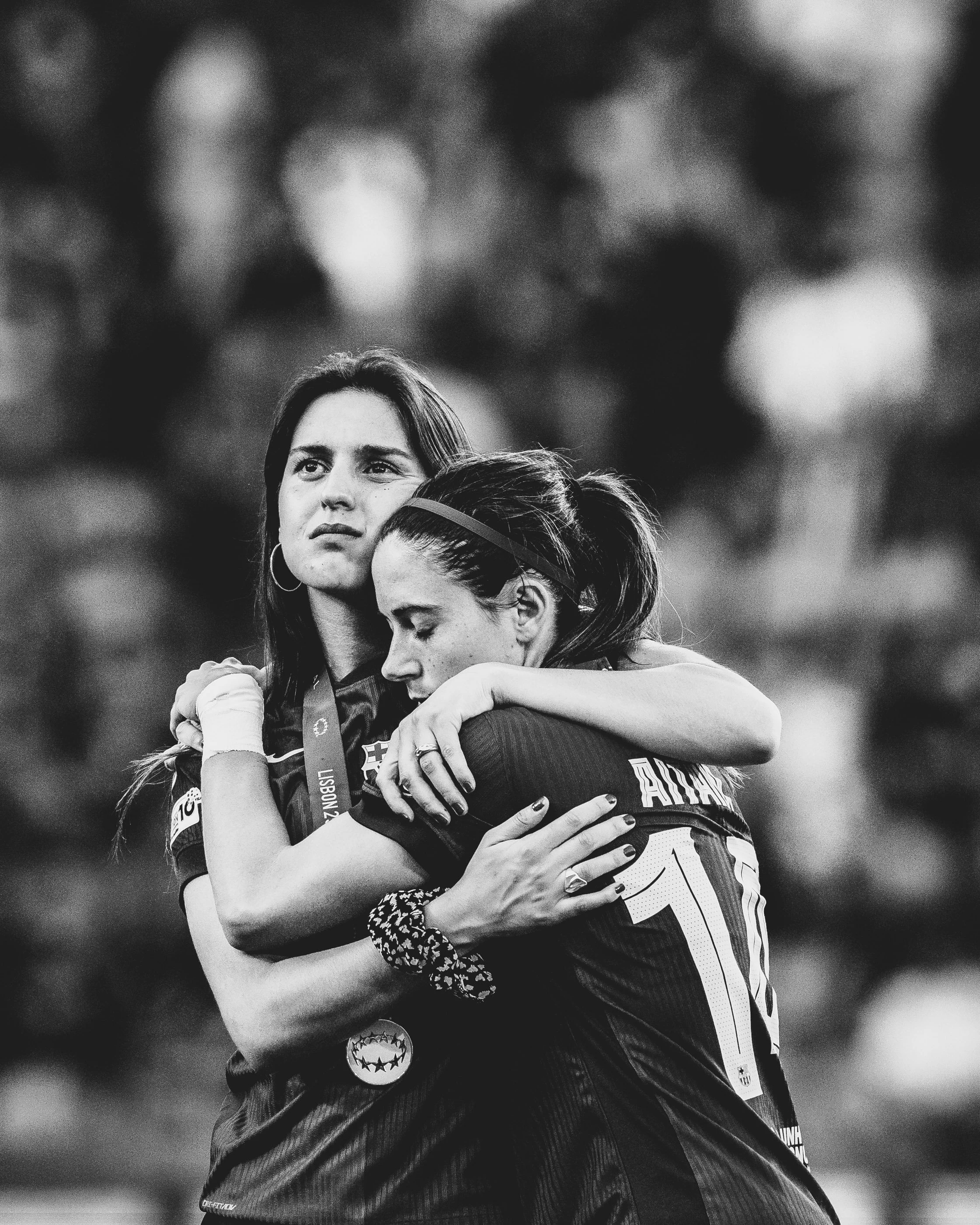 Two women hugging each other, one with closed eyes and the other with an emotional expression, outdoors on a sports field, in black and white.