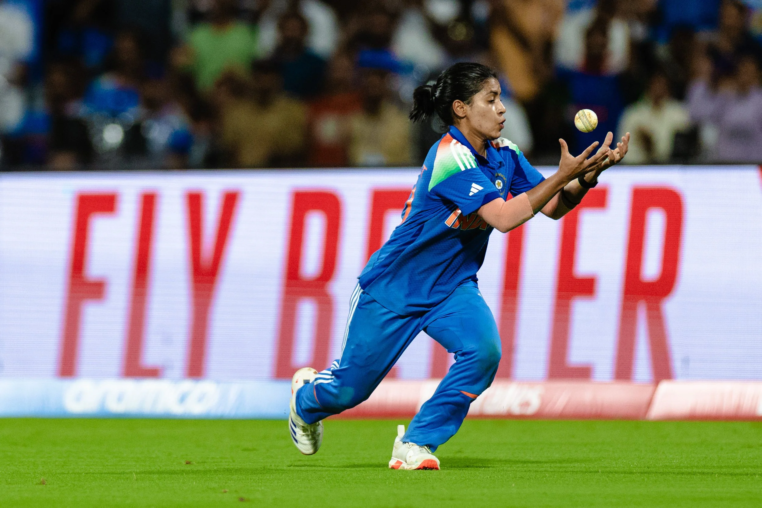A female cricket player in blue jersey and pants runs on a grassy field during a game, with a blurred crowd in the background.