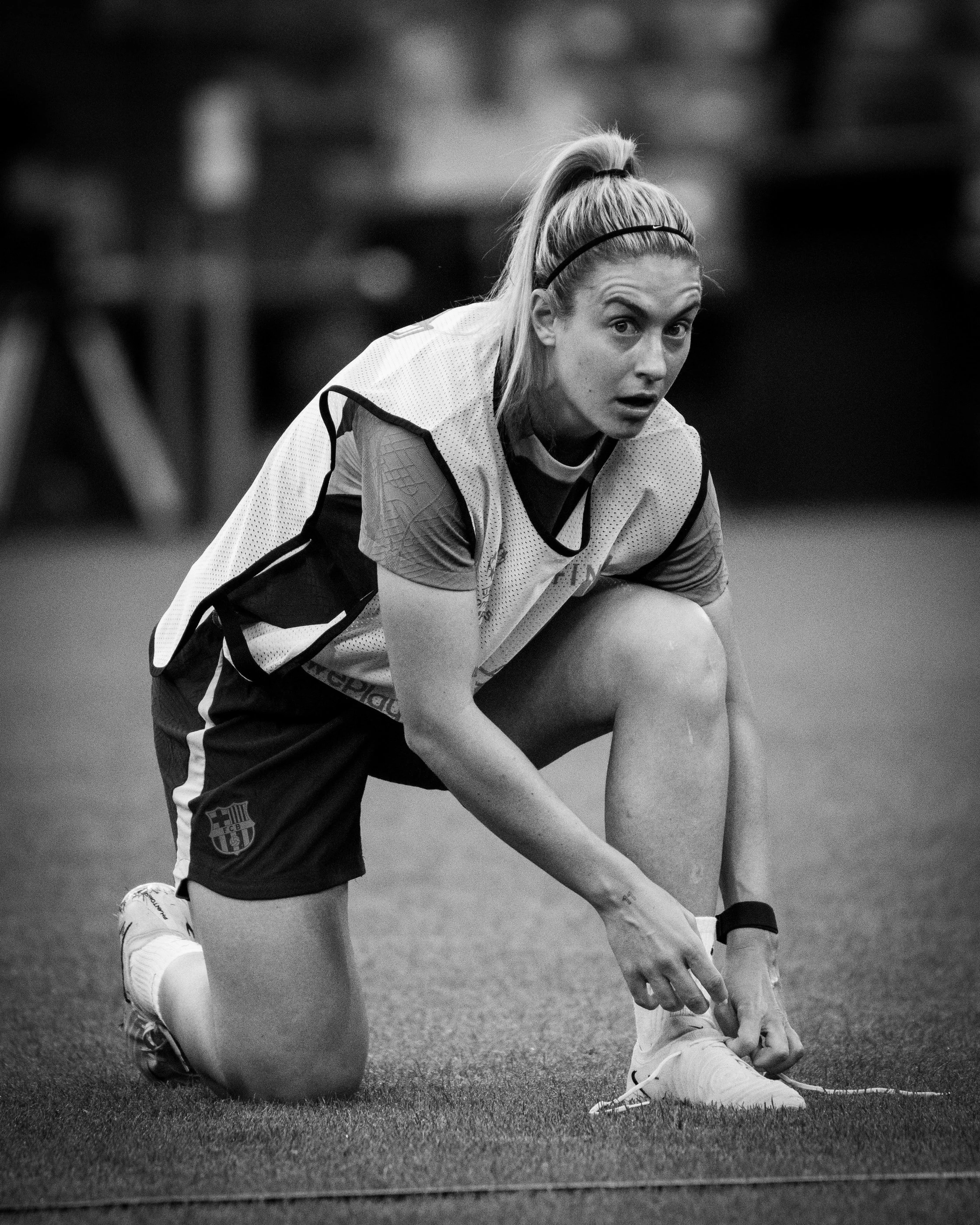 A female athlete is kneeling on a grassy field, tying her shoelace, wearing sports attire including a jersey with a sports emblem, and her blonde hair is tied back with a headband.