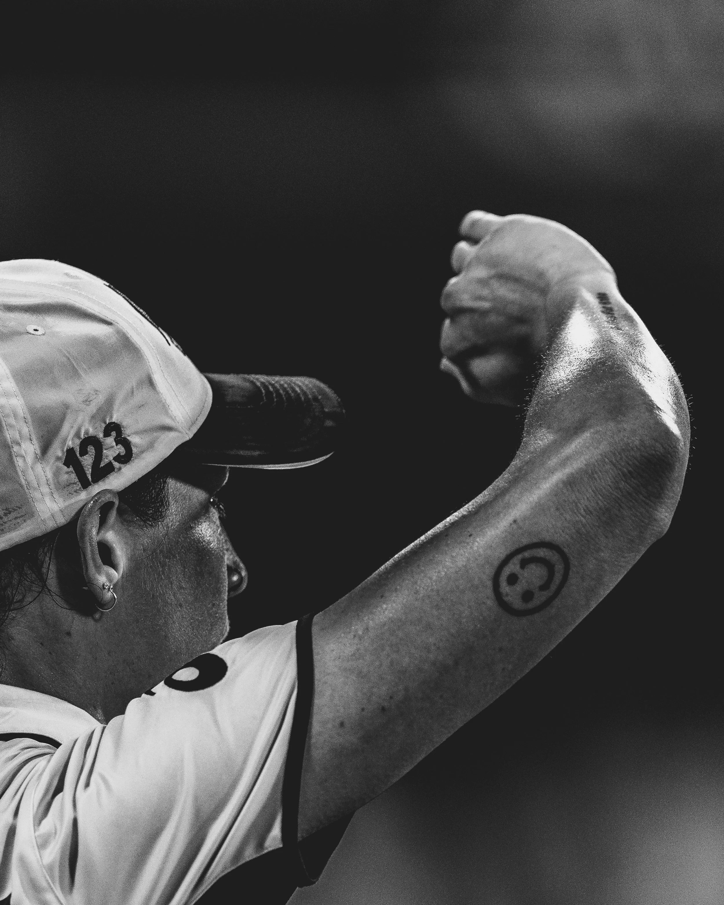 Close-up of a person in a sports jersey with a smiley face tattoo on their arm, resting their head on a surface, with a white sports shoe partially visible in the foreground.