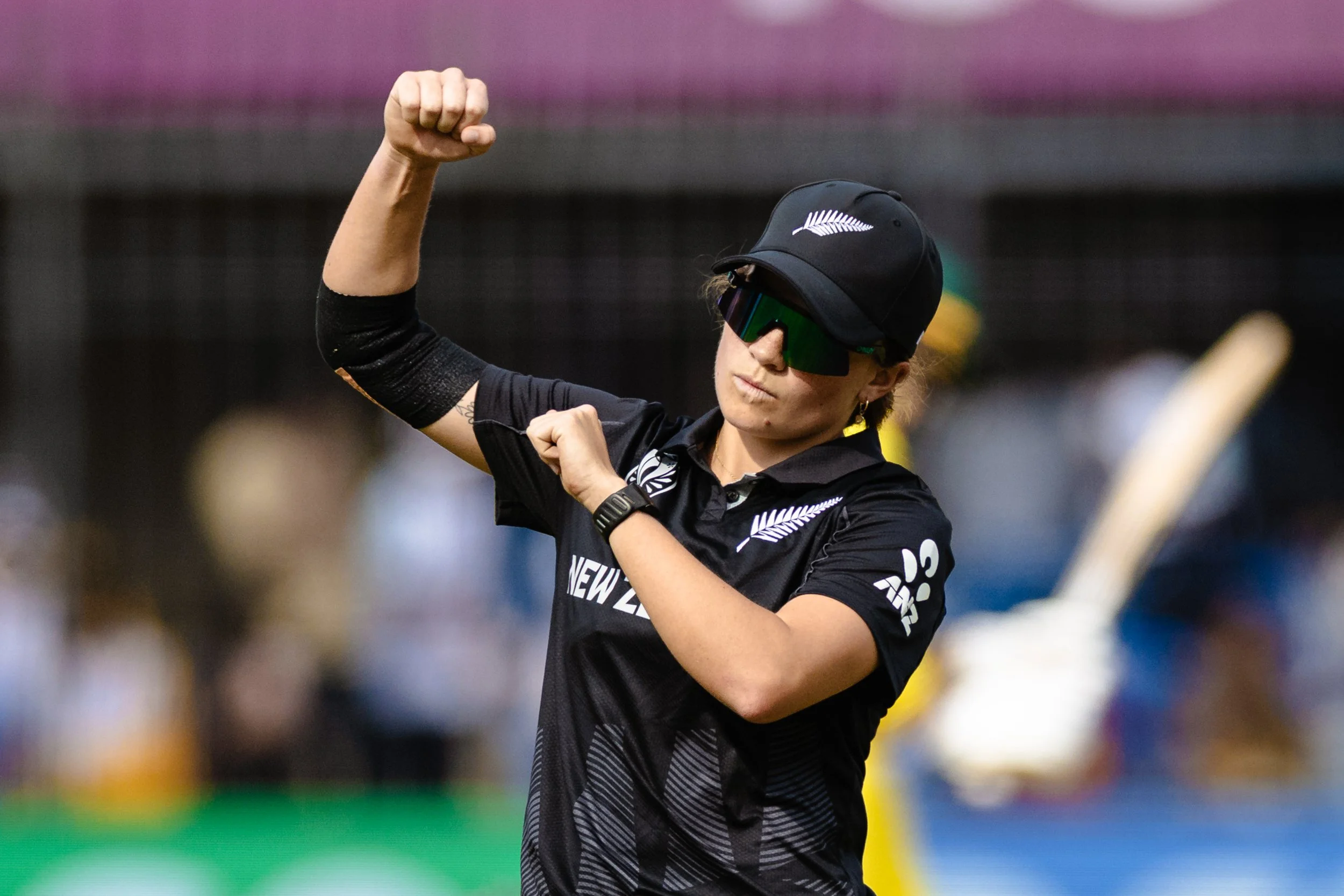 A female cricket player wearing sunglasses, a black hat with the New Zealand fern logo, and a black jersey, is raising her right fist in a victory gesture on the cricket field.