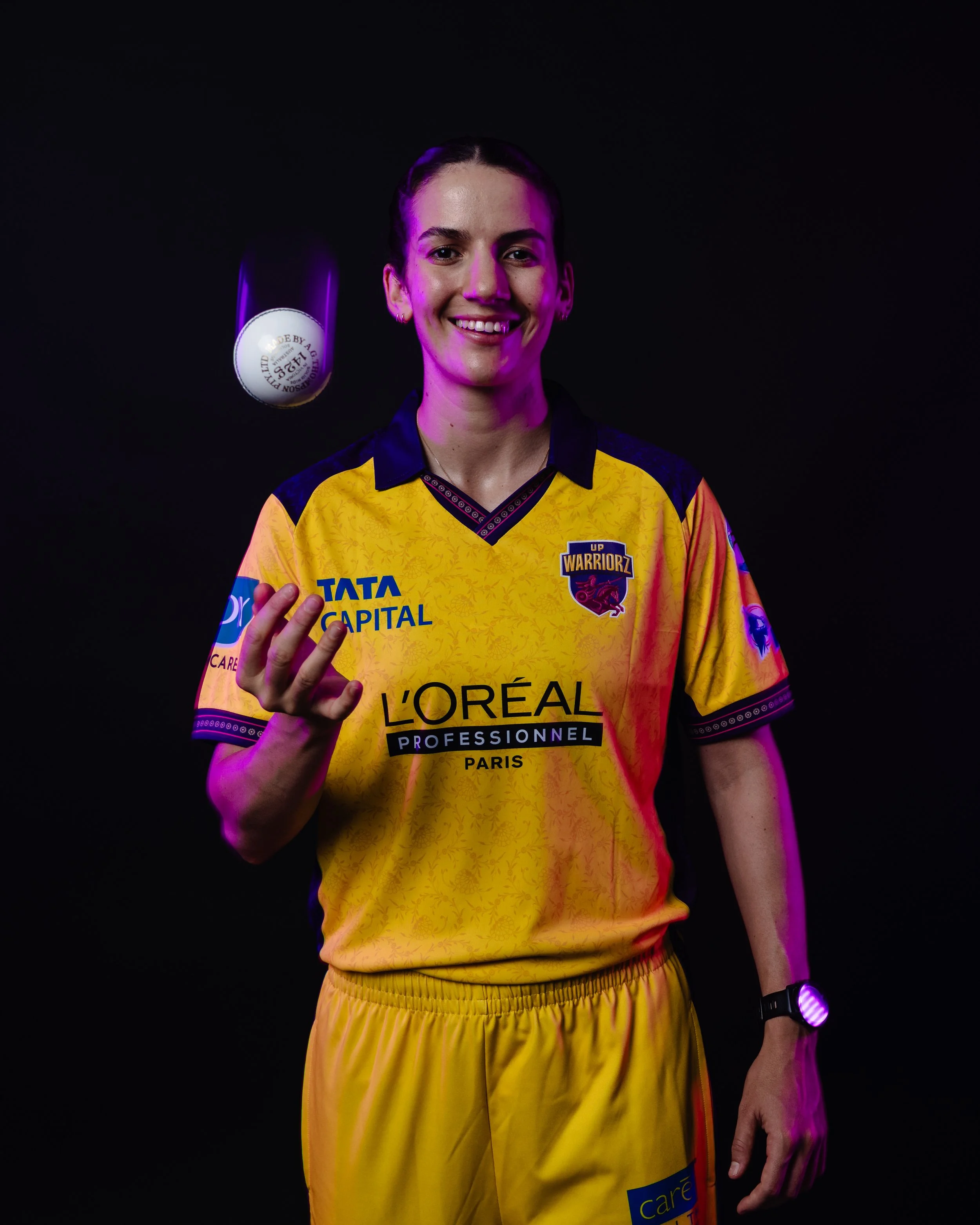 A young female athlete in a yellow sports uniform with the UP Warriors logo, smiling and holding a wristband, against a dark background with purple lighting.