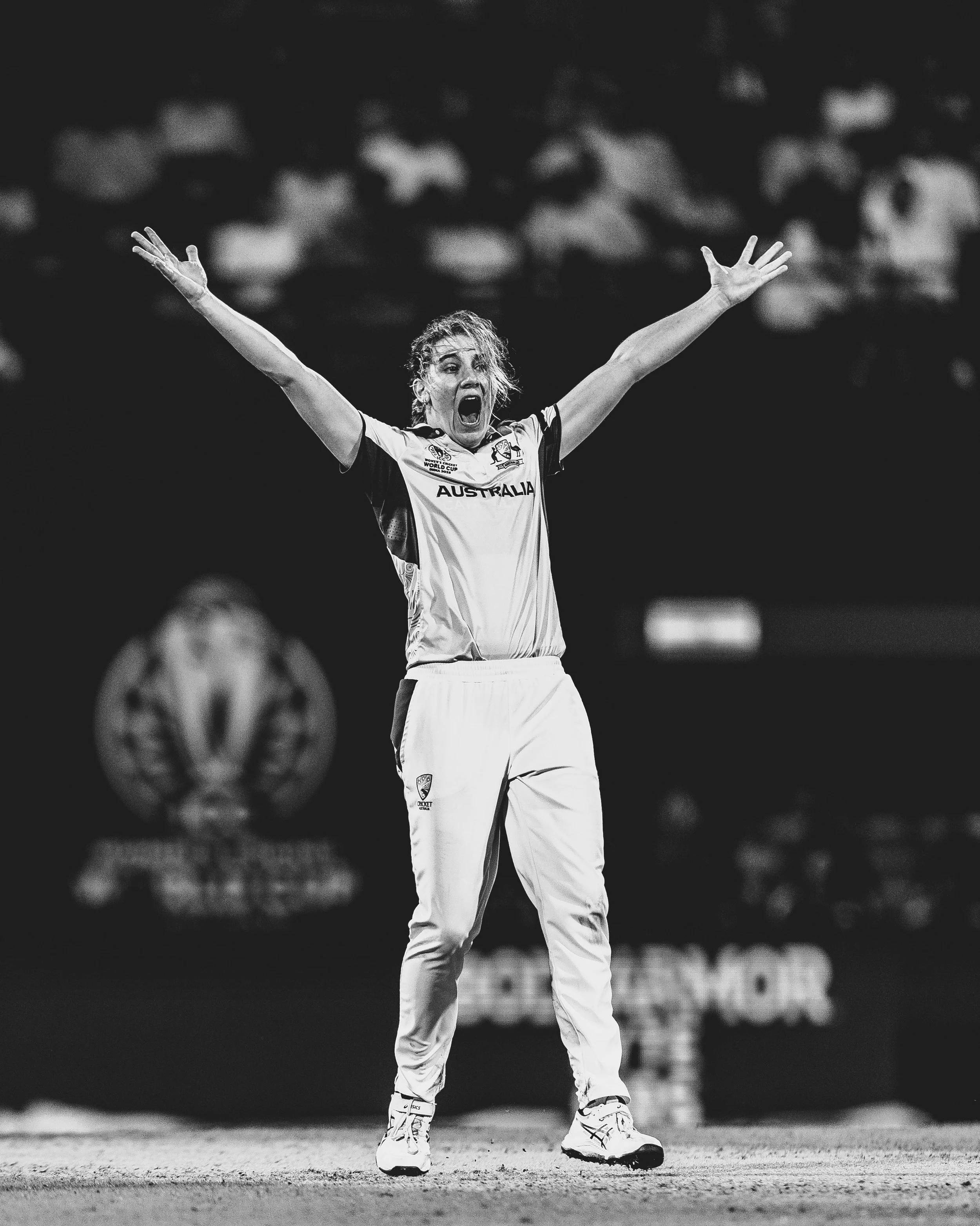 A female cricket player wearing a jersey with 'Australia' written on it, standing on the cricket pitch with her arms raised and mouth open in celebration or shouting.