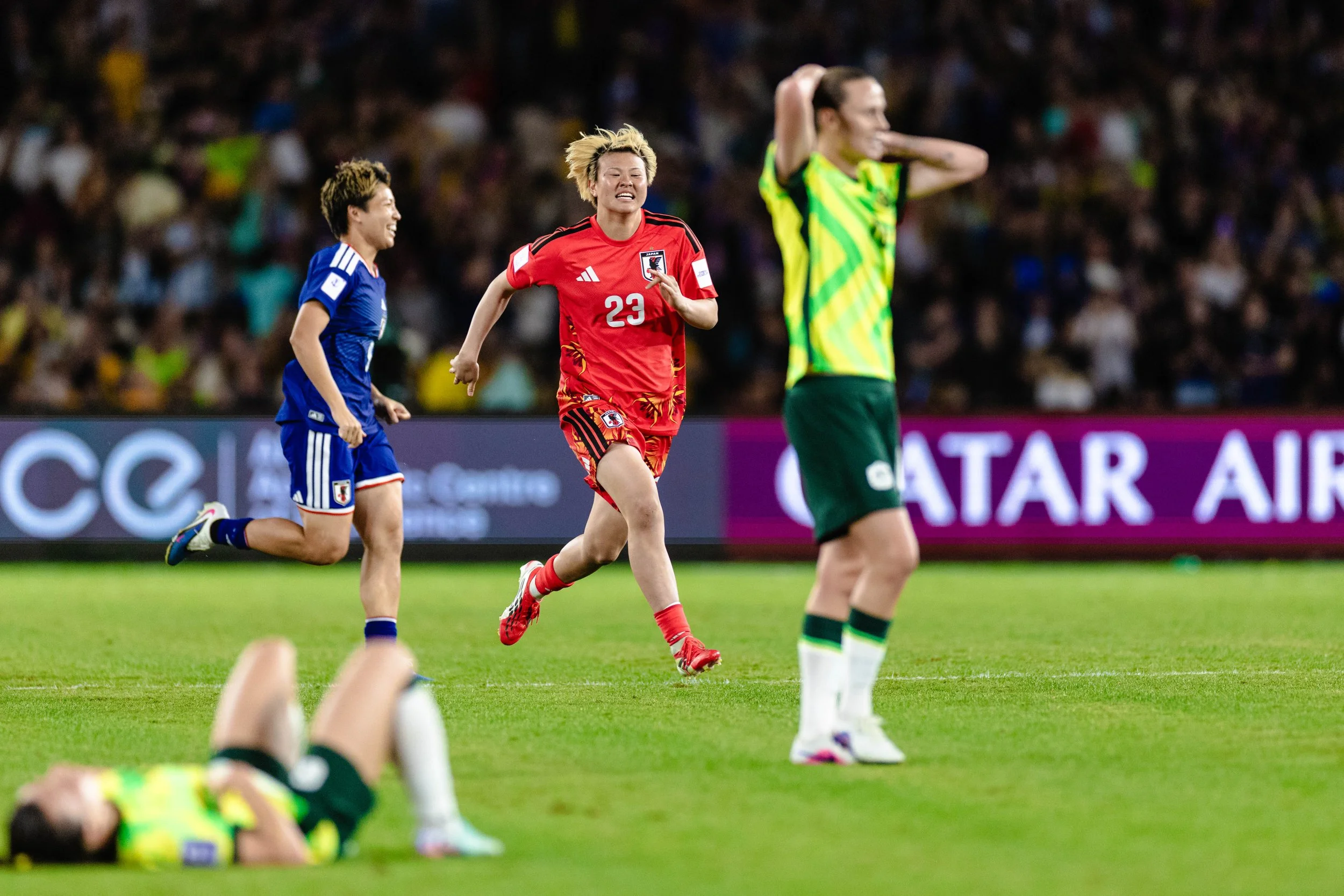A women's soccer match with players running on the field. One player in a red jersey is in the center, with a yellow and green jersey to her right and a blue jersey on the left. The background shows a crowd in the stadium.