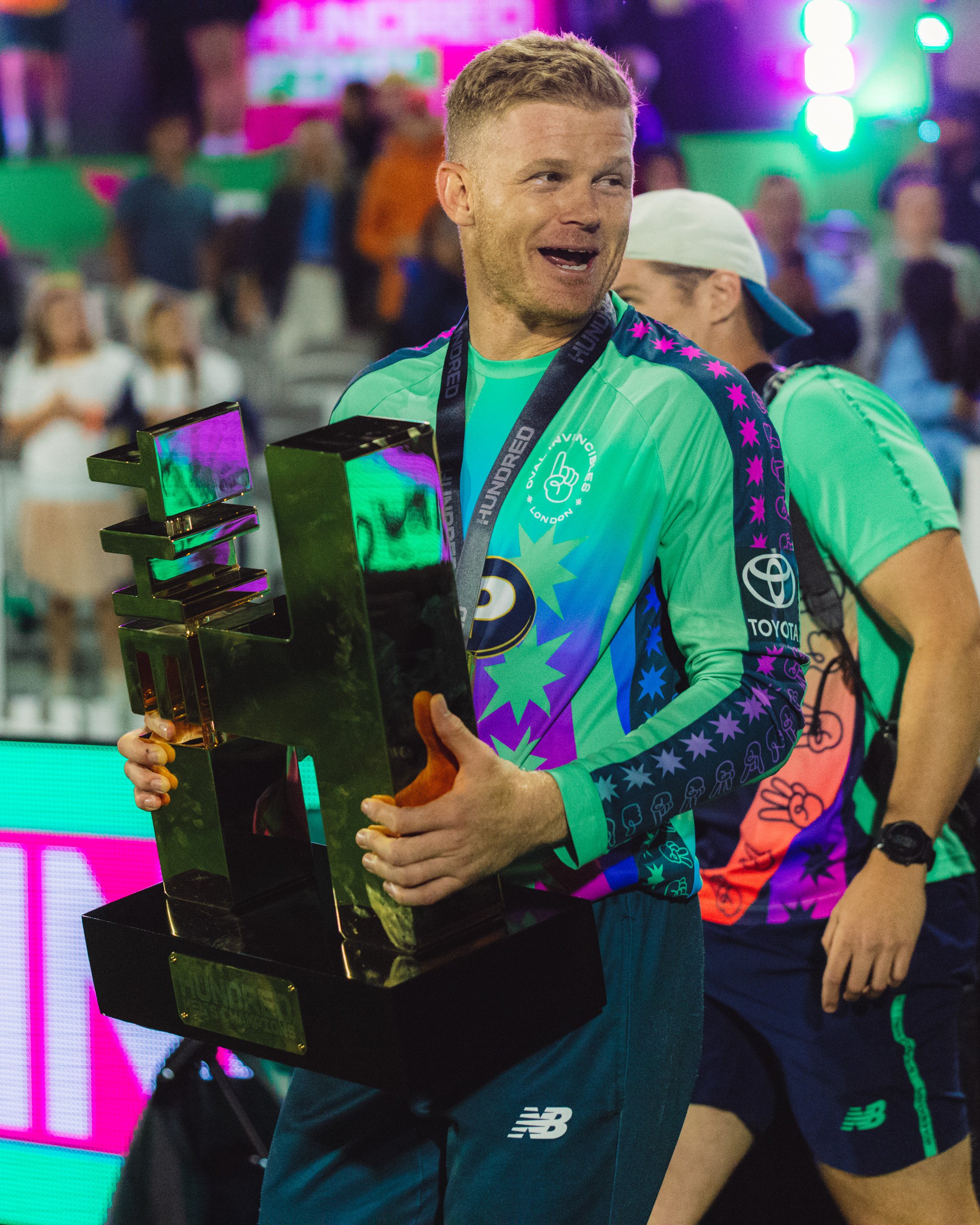 A man wearing a green athletic shirt holding a trophy, smiling after a sports event. Background shows colorful lights and blurred crowd.