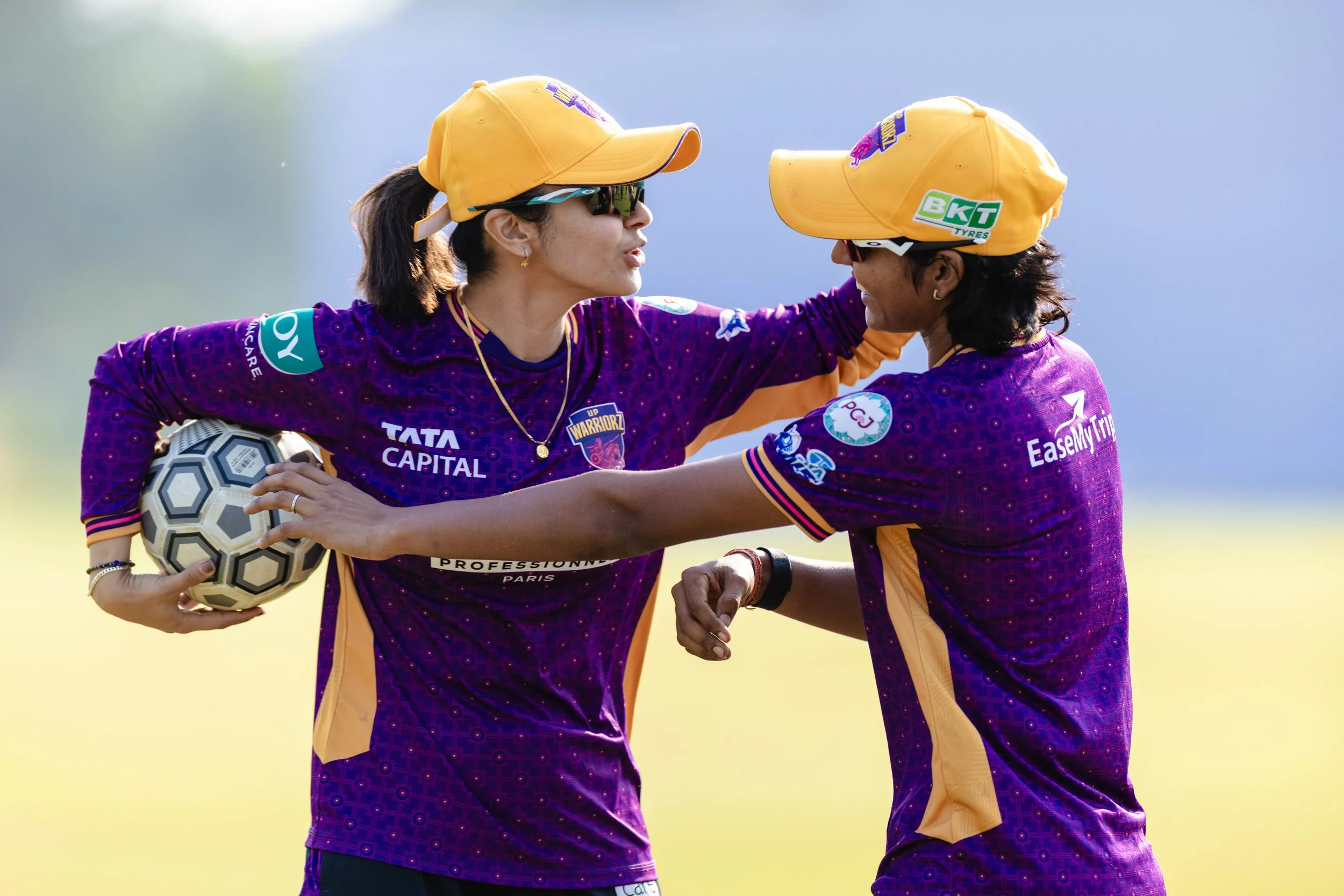Two women in purple and yellow sports uniforms celebrating, one holding a soccer ball, outdoors on a sunny day.