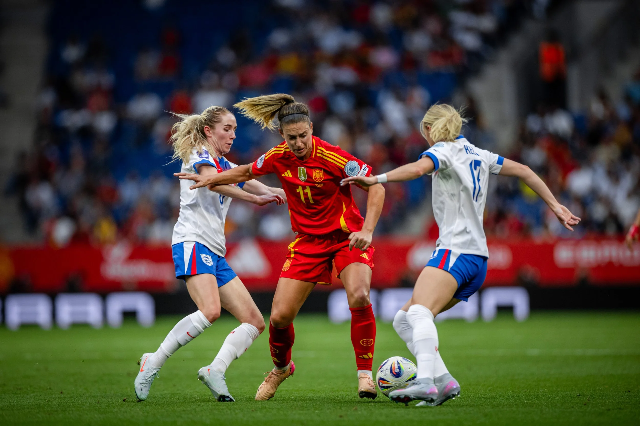Three female soccer players competing for the ball during a game, with two in white jerseys and one in a red jersey.