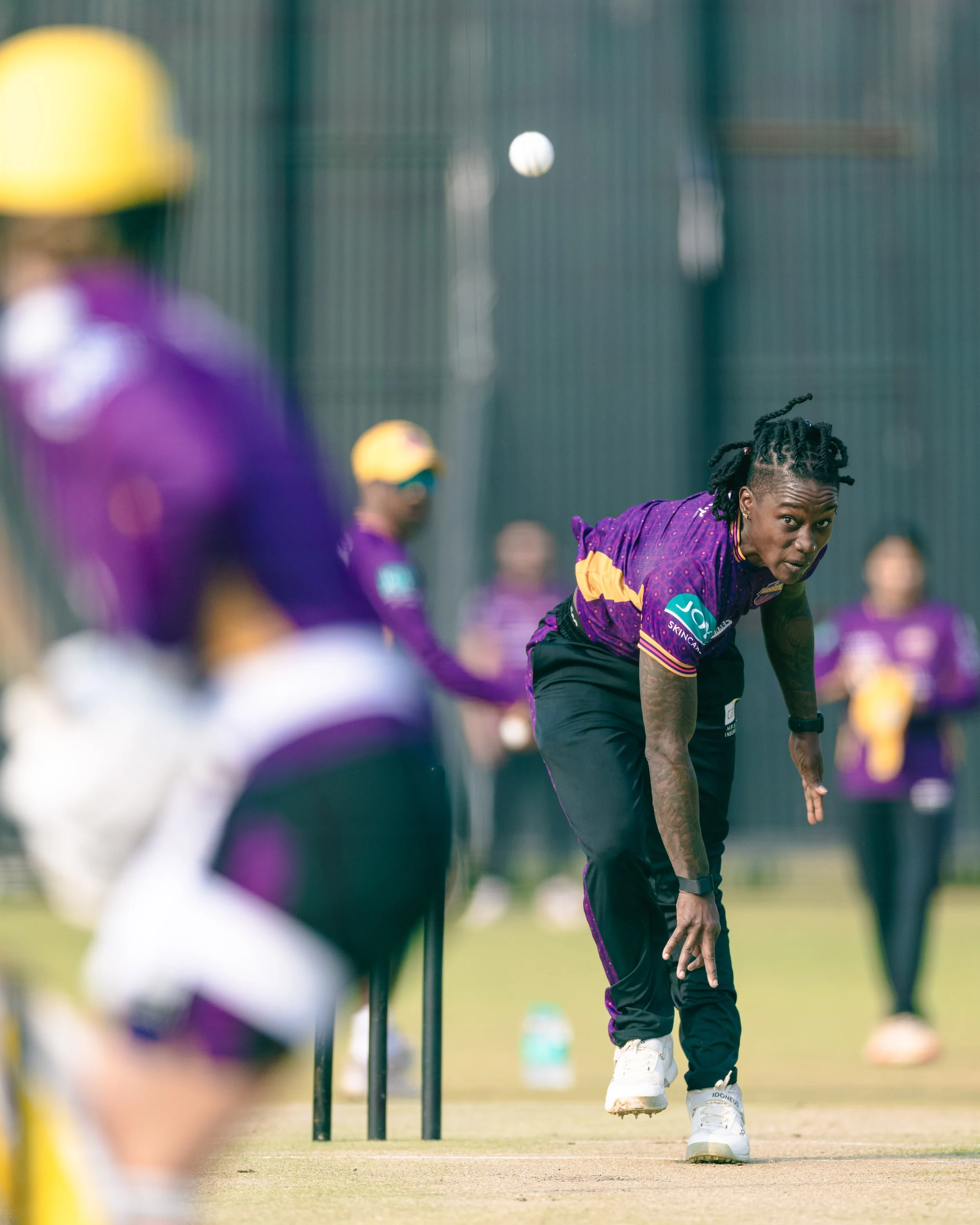 A cricket player in purple and yellow uniform bending down on the field, with other players in purple uniforms in the background, and a white cricket ball mid-air.