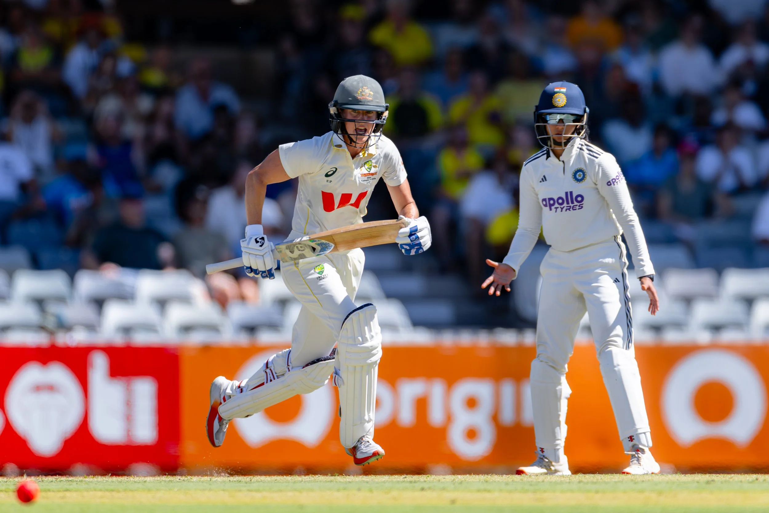 Two female cricket players in white uniforms and helmets running on the field during a match, with a crowd in the background.