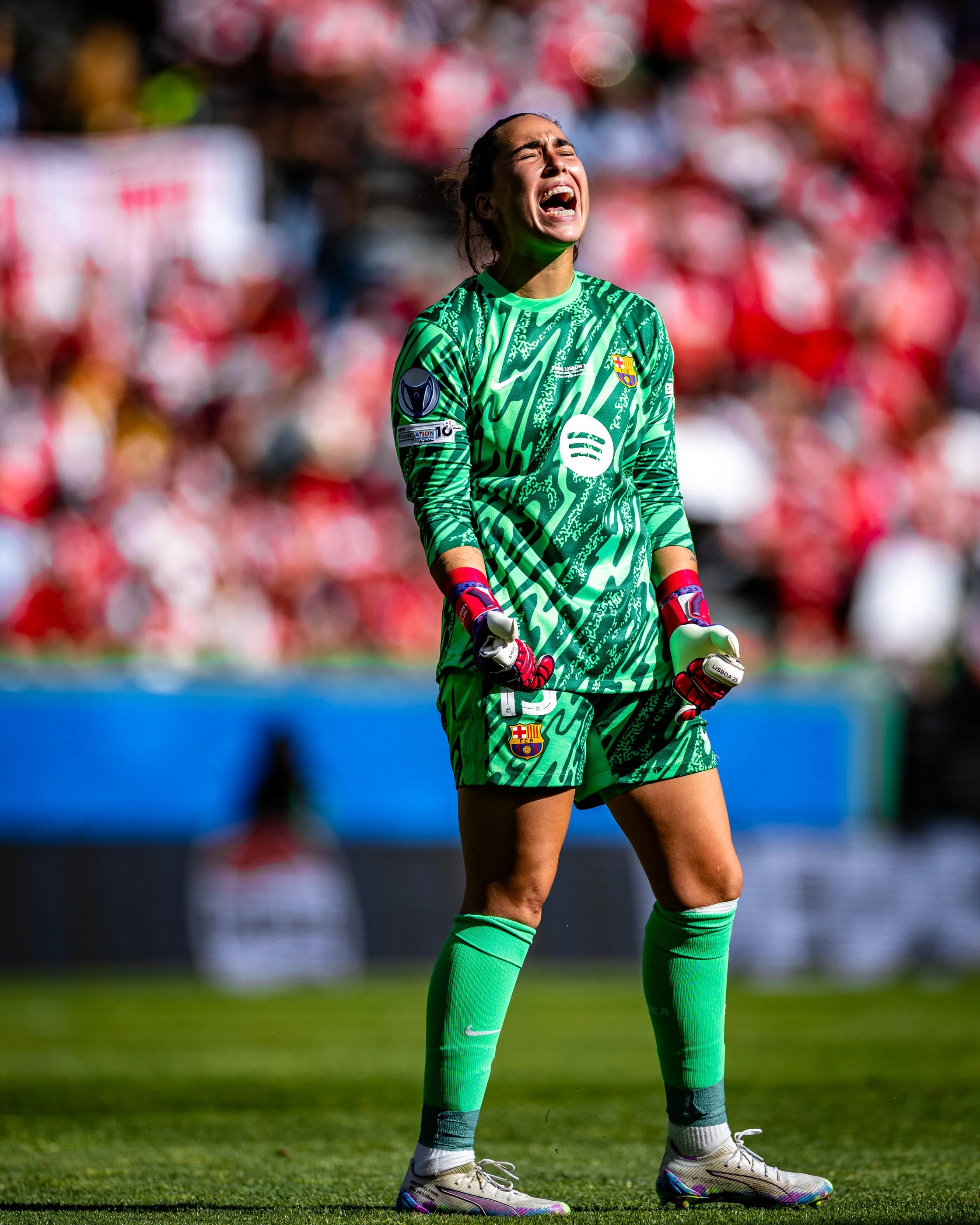 A female soccer goalie wearing a green uniform with the FC Barcelona crest, standing on a field with a disappointed or upset expression, with a blurred crowd in the background.