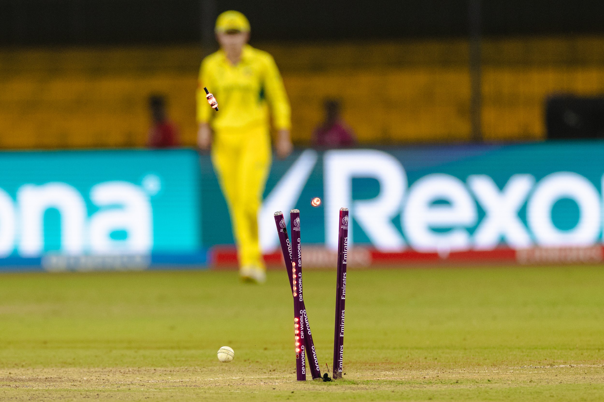 Cricket ball hitting stumps during a match at night, with a player in a yellow uniform in the background.
