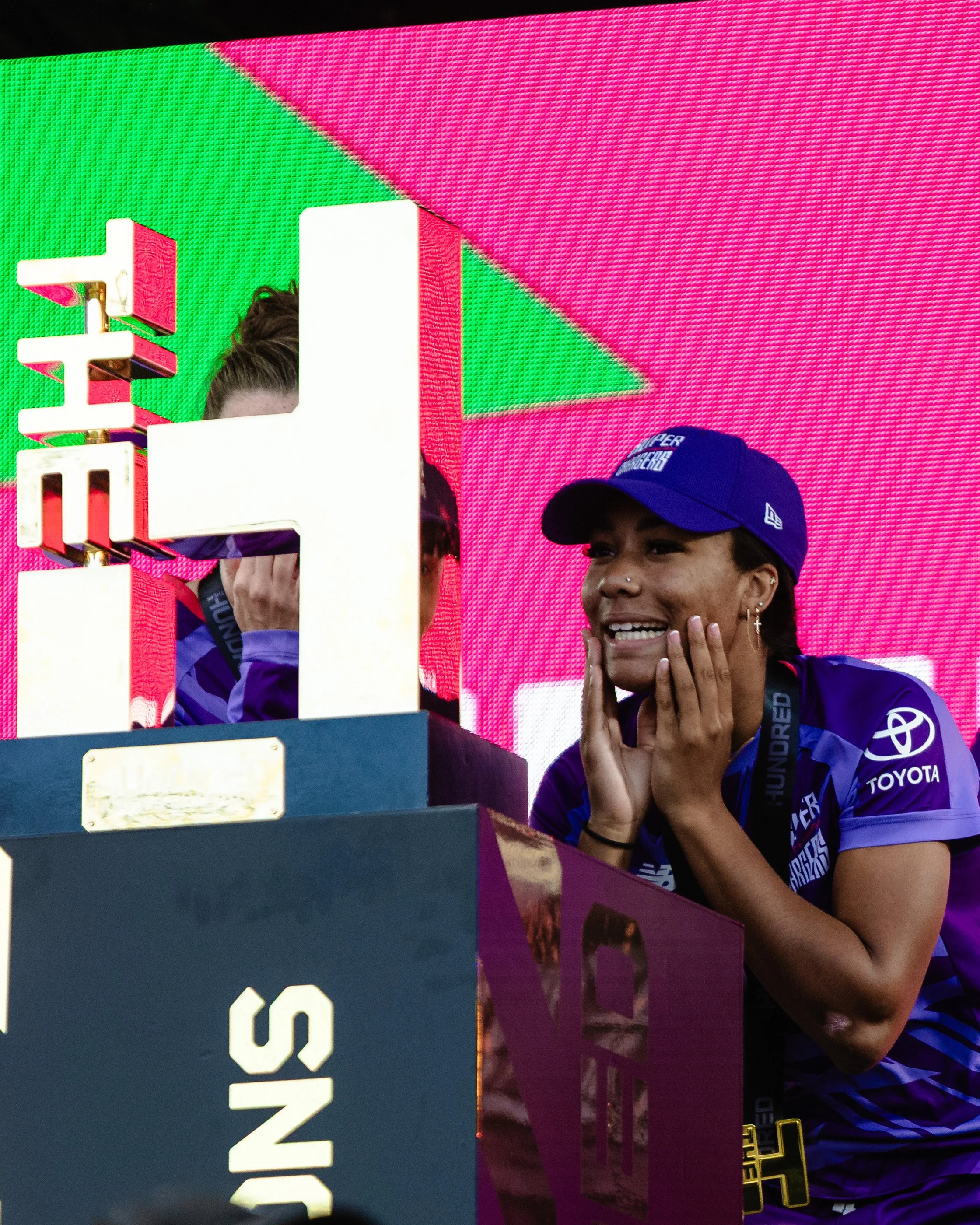 Person smiling and holding hands to face, wearing a purple cap and purple jacket, with a large digital screen in the background displaying pink, green, and purple colors.