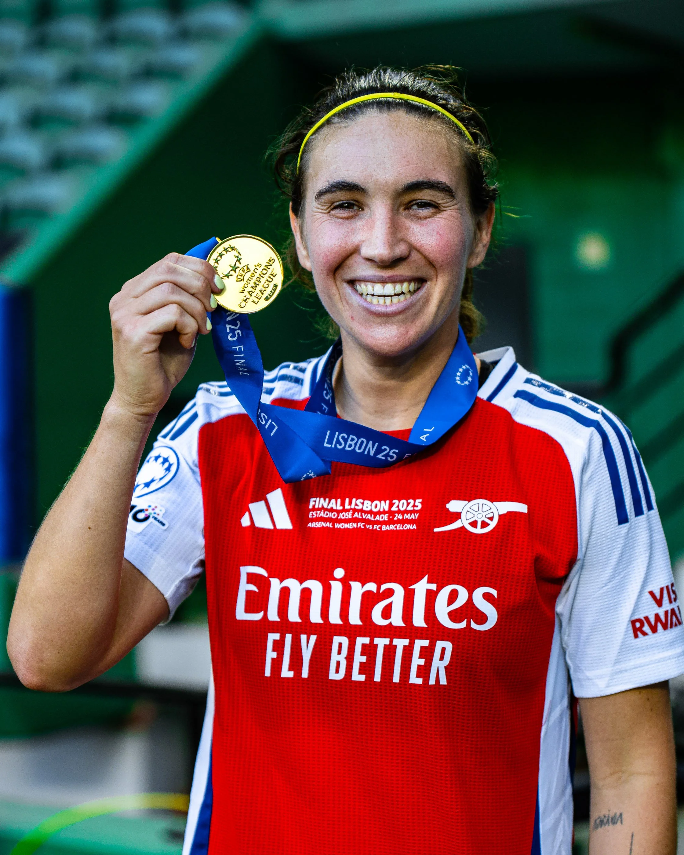 Female athlete holding a gold medal at a sports event, wearing a red and white jersey with blue accents and a blue ribbon around her neck. She is smiling, with a tattoo visible on her right arm. The background shows a green structure.