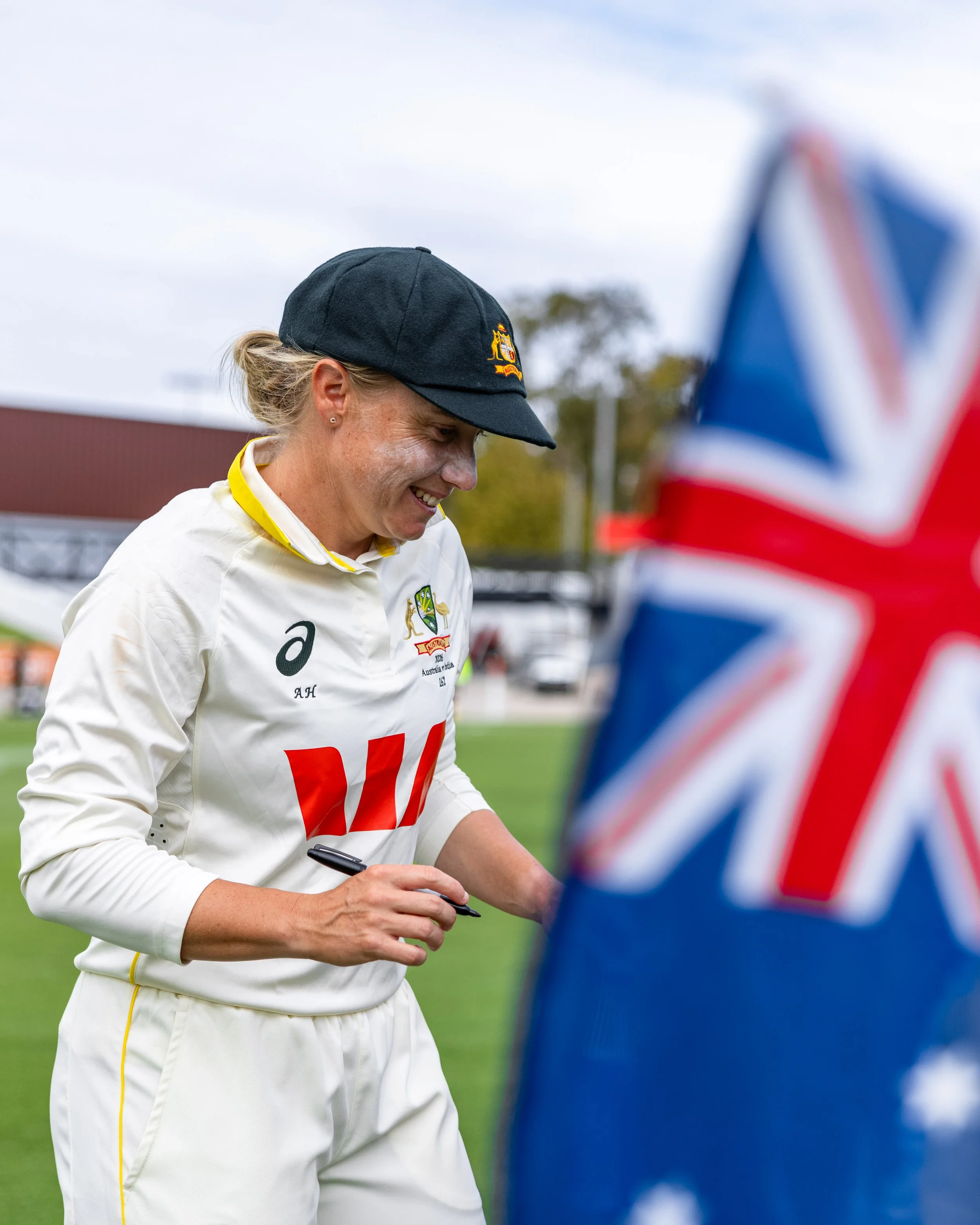 A smiling woman in a white sports uniform and navy cap signing autographs or writing on something, with a blurred flag in the foreground and a cloudy sky in the background.