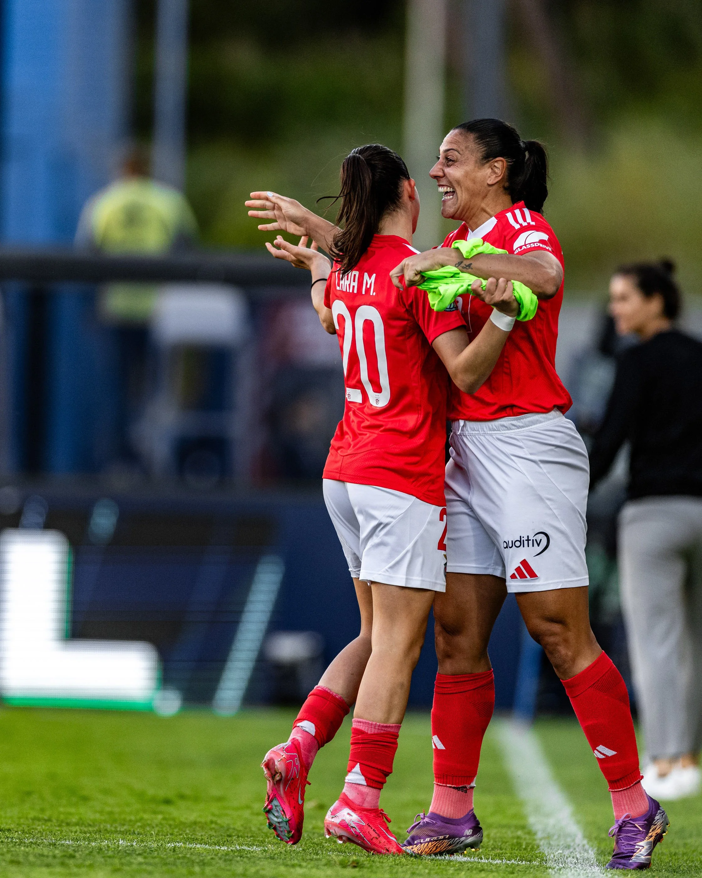 Two female soccer players celebrating on the field, hugging and smiling, wearing red jerseys and shorts with sports shoes, with a blurred background of a soccer field and a person in yellow in the distance.