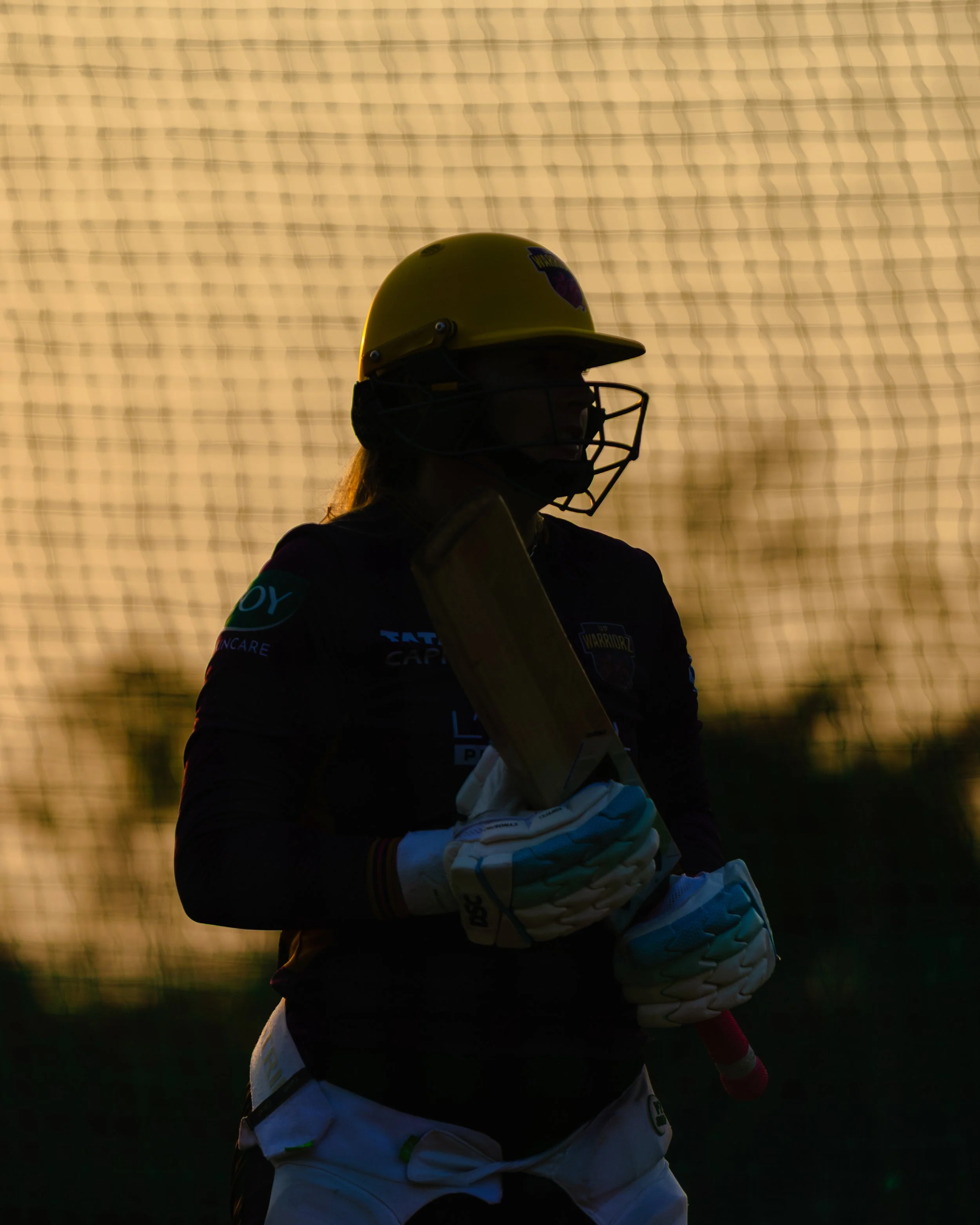 Silhouette of a cricket player wearing a yellow helmet with a faceguard, holding a cricket bat, with a wire fence in the background during sunset or sunrise.