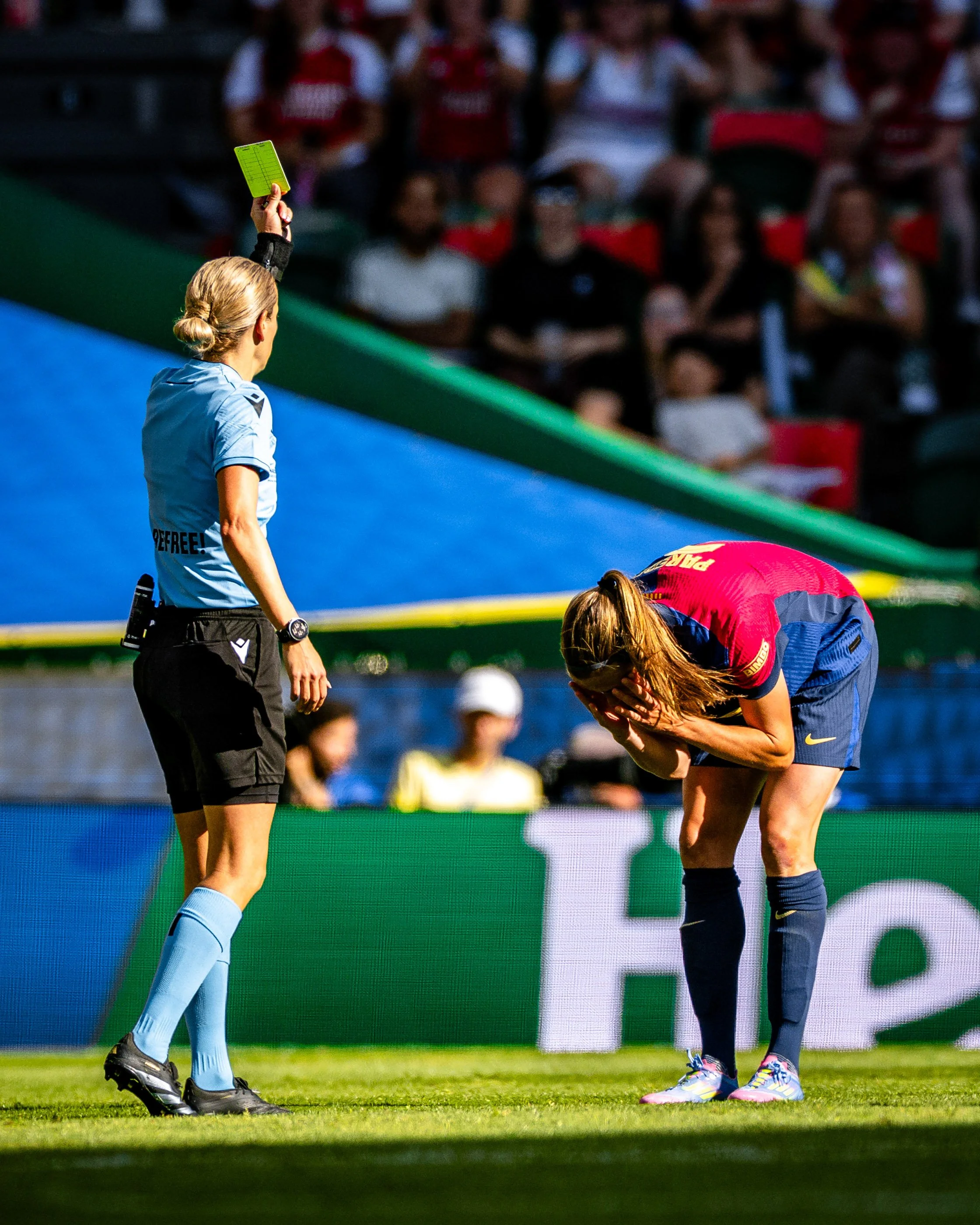 A female soccer player in a red and blue uniform bends over on the field, while the referee, a woman with blonde hair tied back, holds up a yellow card during a match. The stands are filled with seated spectators.