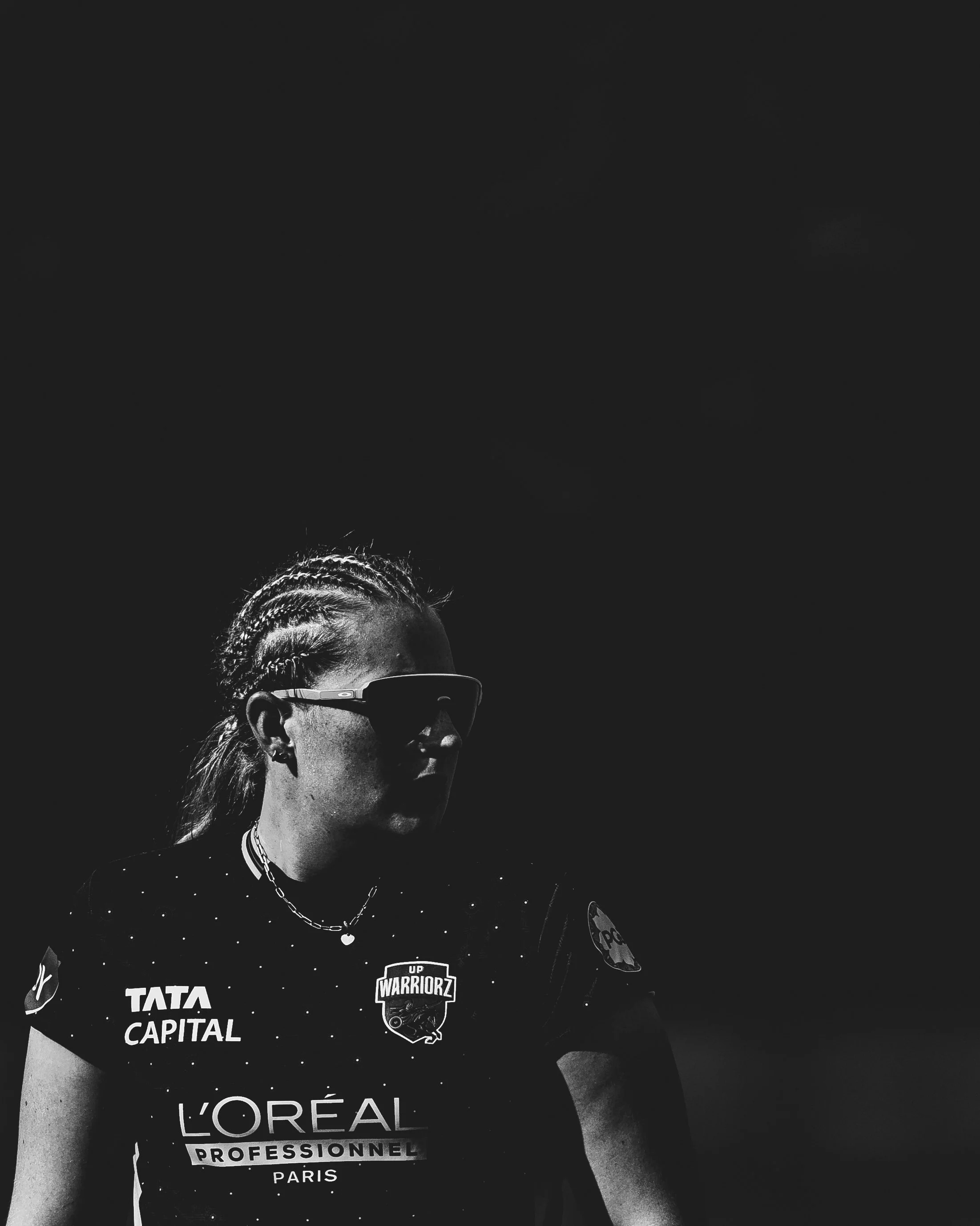 A woman with braided hair wearing sunglasses, a black t-shirt with sponsor logos, and a silver chain necklace, standing against a dark background.