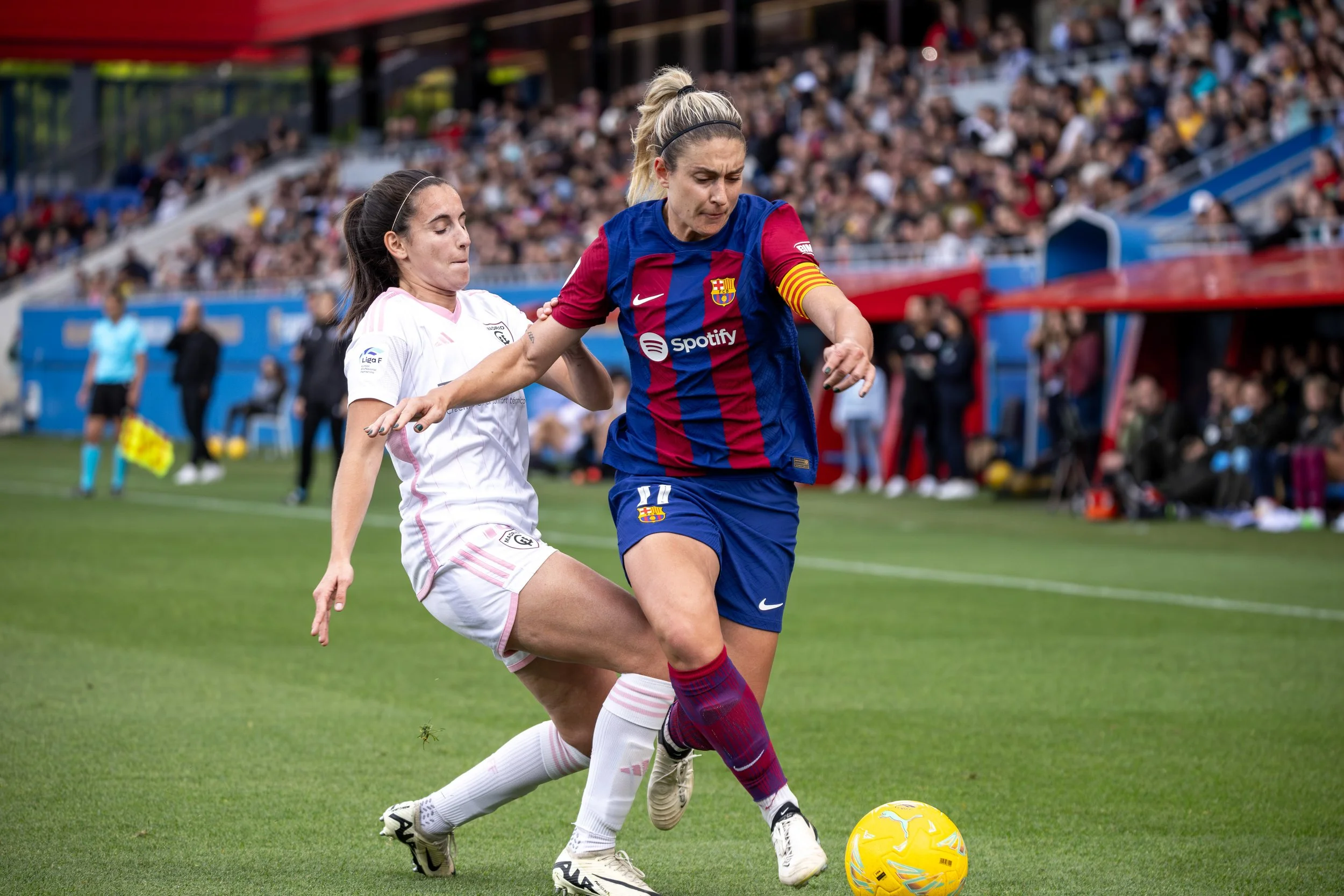 Two women playing soccer, one in a white jersey and the other in a FC Barcelona jersey, competing for the ball on a soccer field with a crowd in the background.