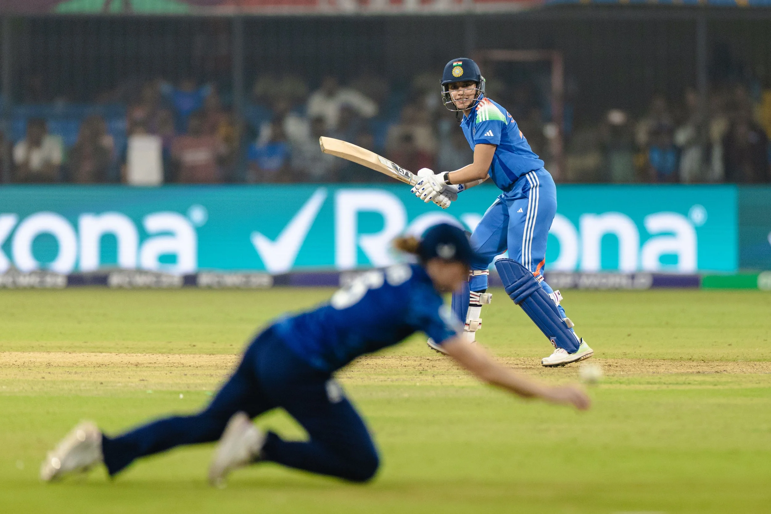 A cricket player in blue uniform and helmet preparing to hit the ball, with a fielder diving on the ground in the foreground during a match.